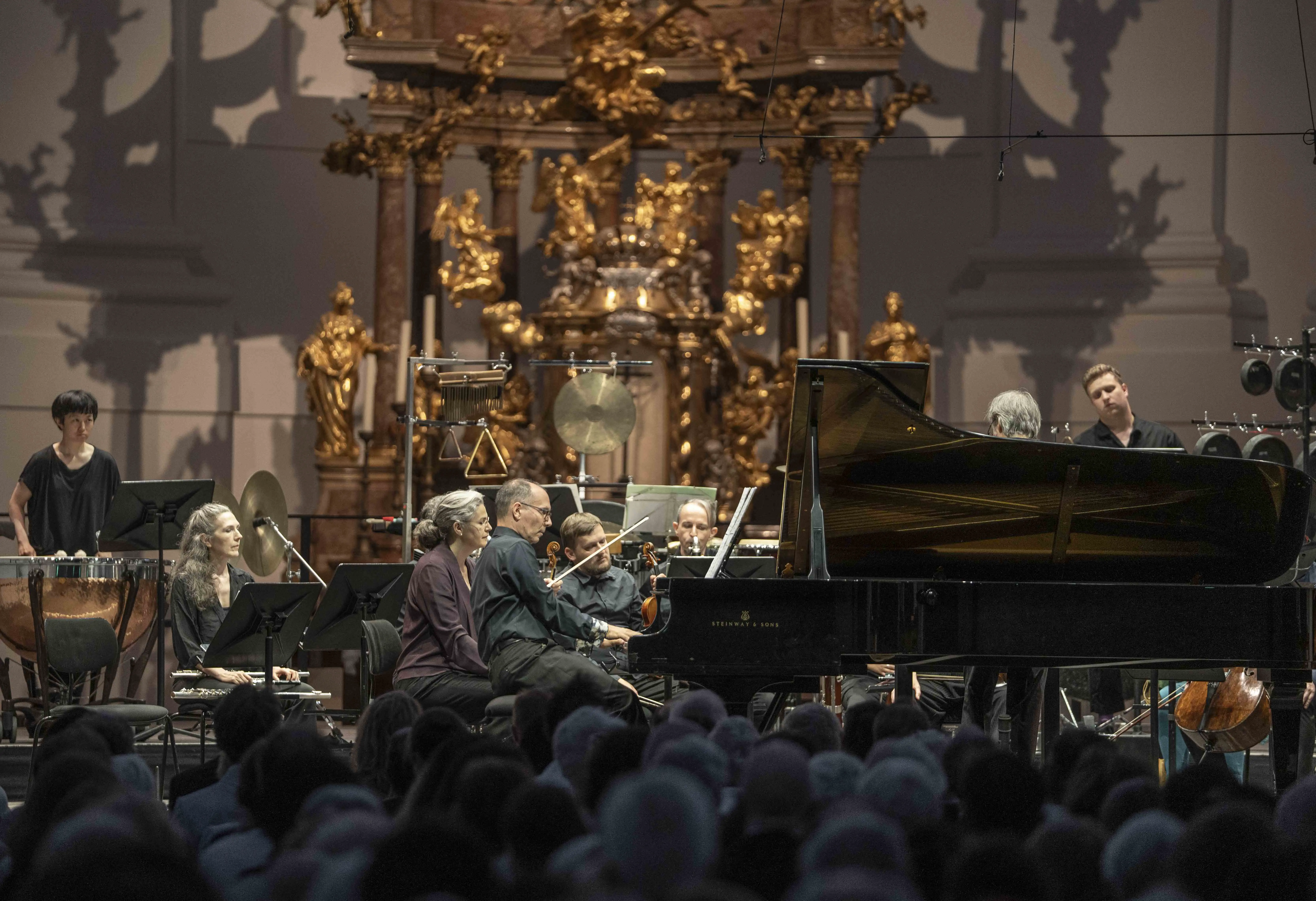 Musicians performing 'Tenebrae Responsoria' at the 2025 Salzburg Festival in the baroque Kollegienkirche, with grand piano, percussion, and strings set against a gilded altar backdrop.