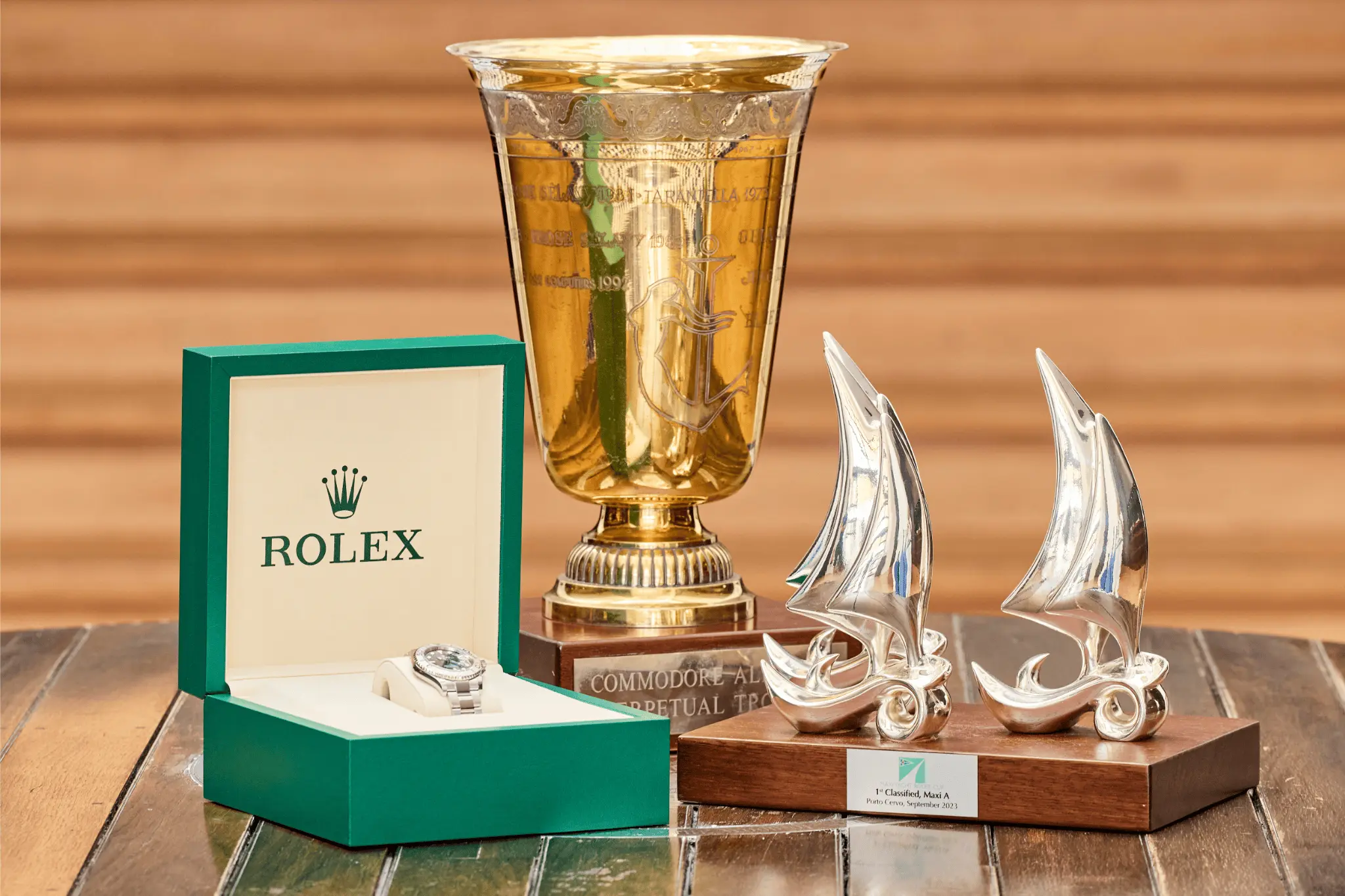 Close-up of the Rolex timepiece and sailing trophies awarded at the Maxi Yacht Rolex Cup 2025. The display includes a green Rolex watch box, a golden perpetual trophy, and two silver sailboat-shaped trophies on a wooden table.