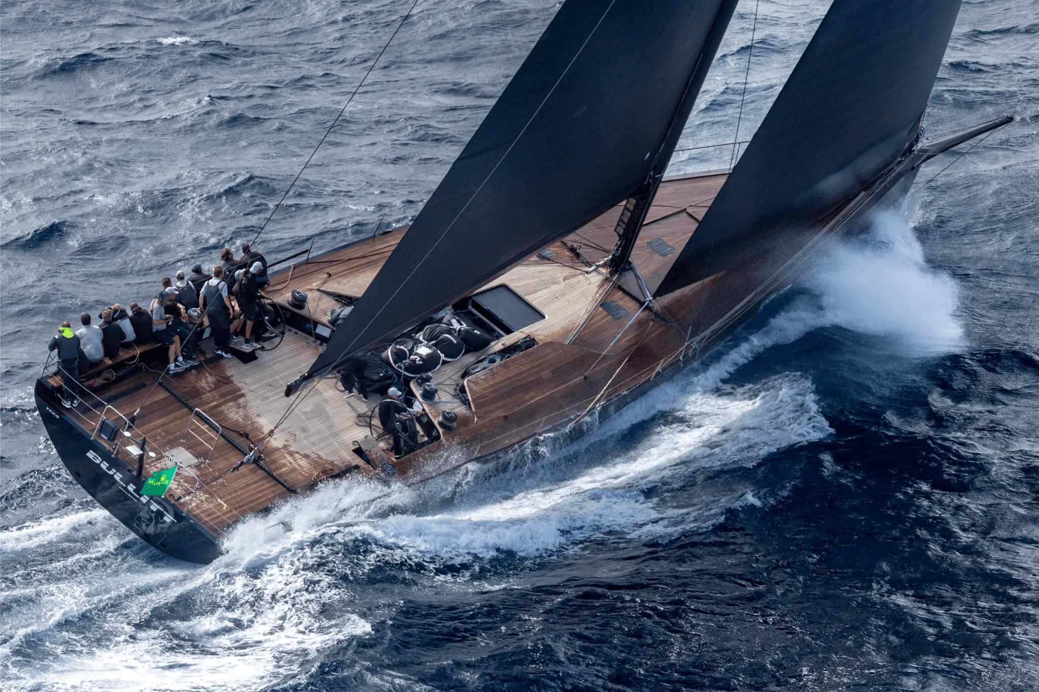 Aerial view of the yacht Bullitt slicing through the waves with full sails during the Maxi Yacht Rolex Cup 2025. The crew is gathered on deck as the sleek, wooden-decked vessel powers forward in deep blue seas.