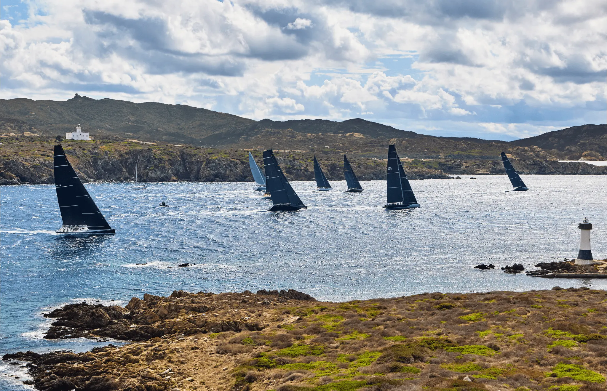 Fleet of high-performance maxi yachts racing along the dramatic coastline of Sardinia's Maddalena Archipelago, with rocky shores and a lighthouse in the foreground during the 2025 Maxi Yacht Rolex Cup.
