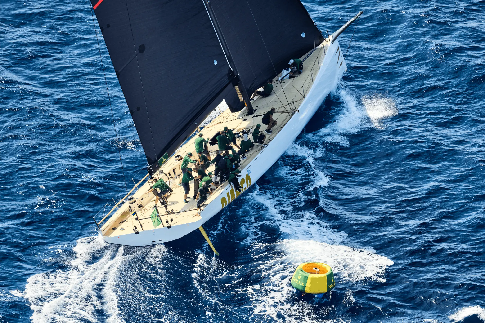 The yacht Django 7X, with crew in green uniforms, rounding a yellow and green Rolex-marked buoy during the Maxi Yacht Rolex Cup 2025. The sleek white hull cuts through the deep blue sea as the team works in perfect coordination.