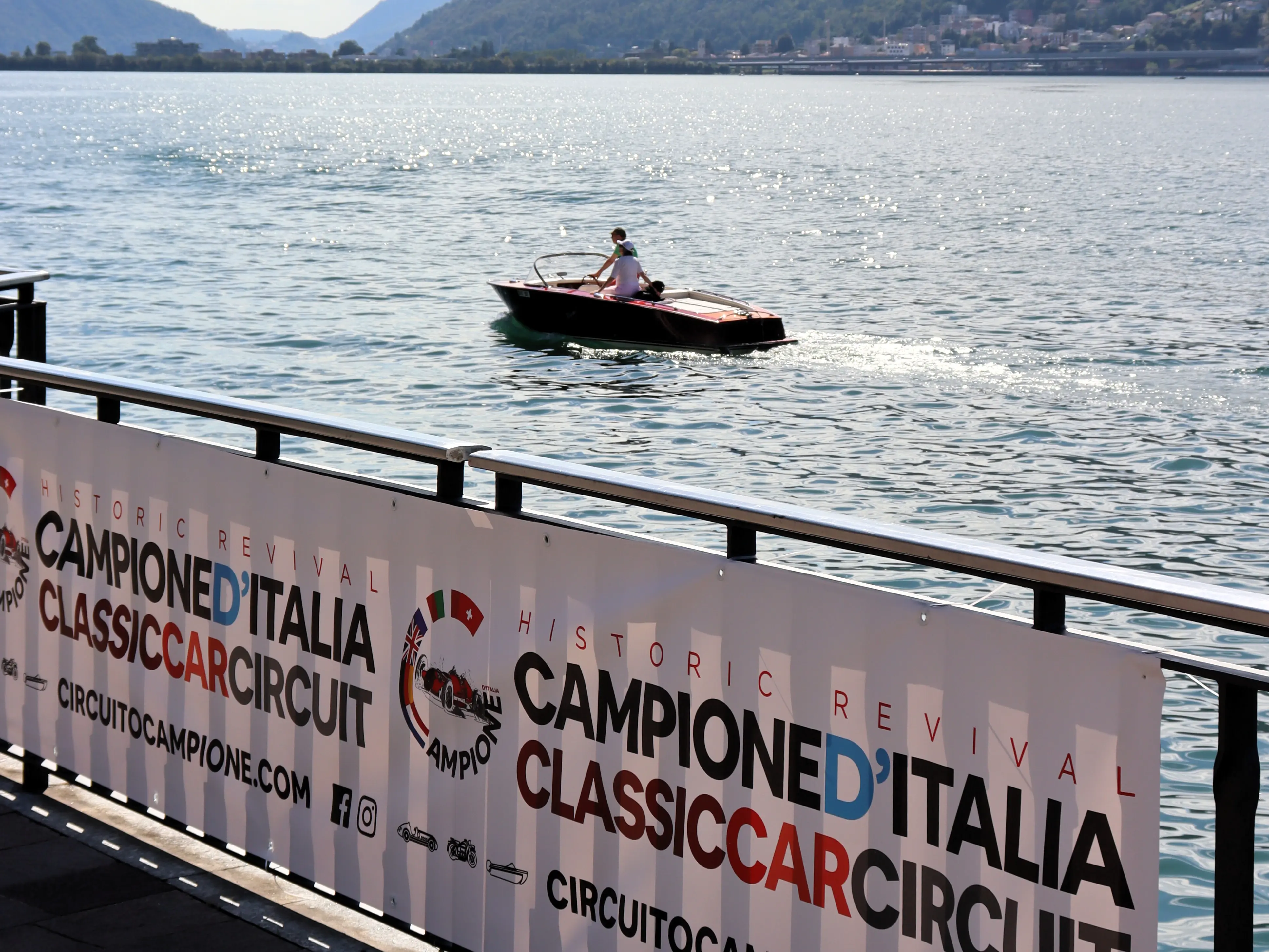 A vintage Riva speedboat glides across Lake Lugano behind a banner promoting the Campione d’Italia Classic Car Circuit, with scenic mountains in the distance.