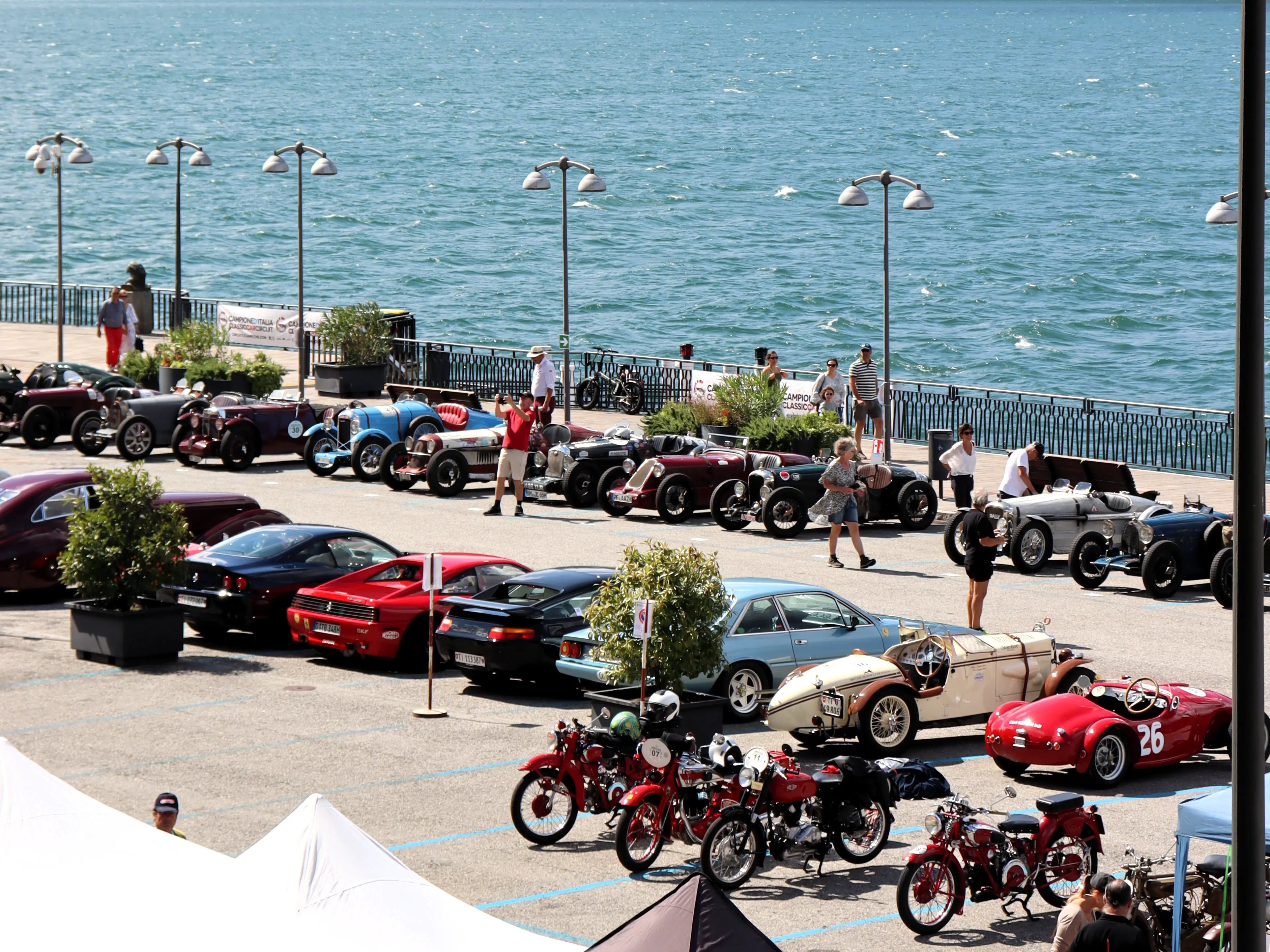 Rows of pre-war and post-war cars and motorcycles are parked beside Lake Lugano, with spectators admiring the vintage machines under the sun.