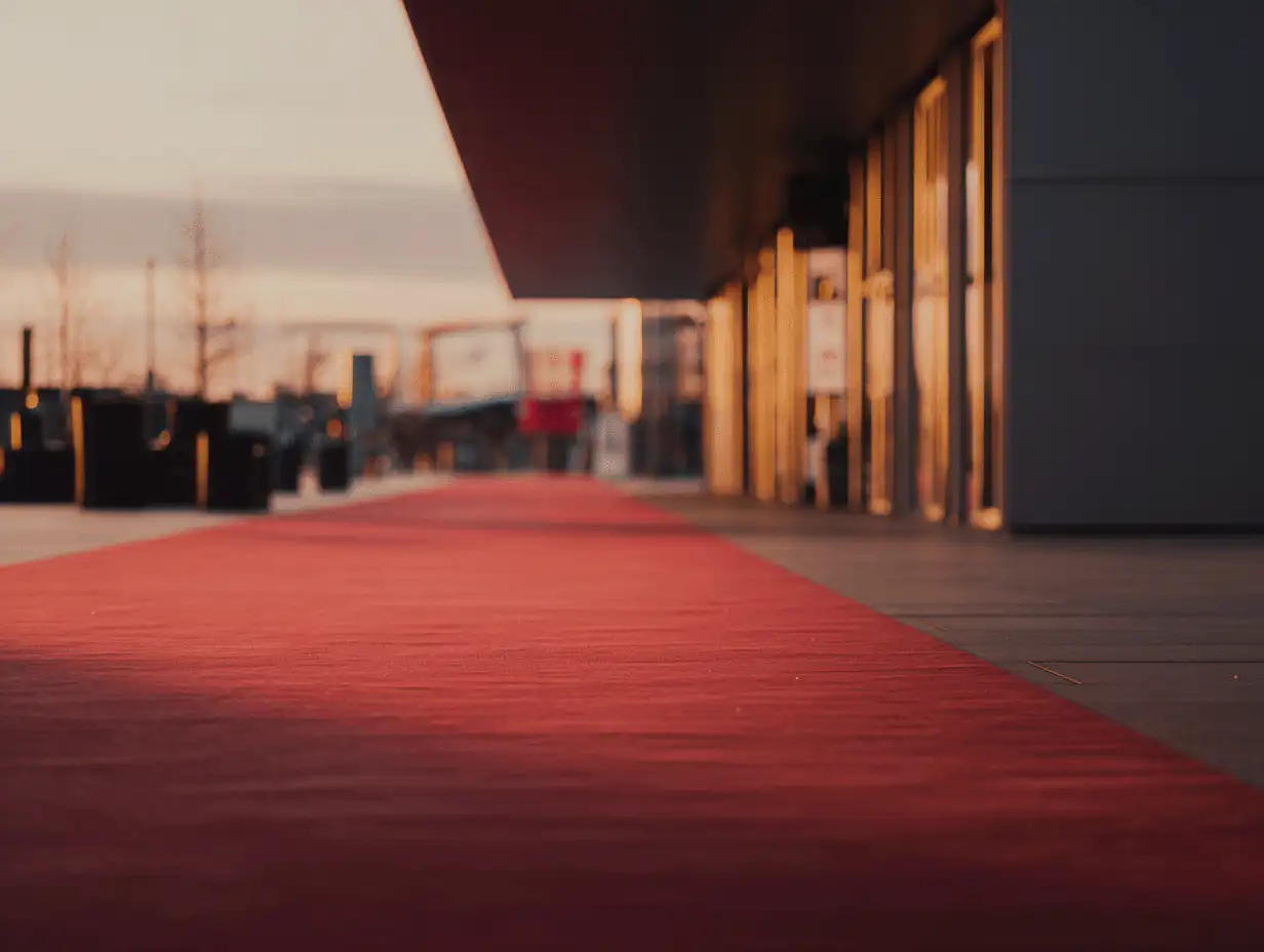 A glowing red carpet leads into a glass-walled venue at dusk, evoking the anticipation and allure of cinematic premieres and cultural spotlight.