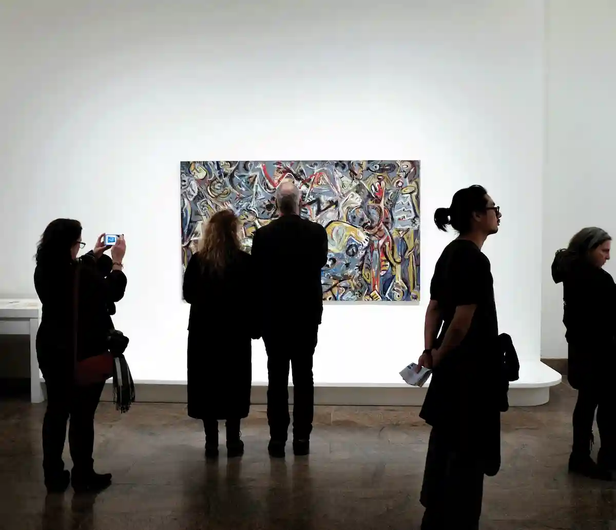 Visitors in a modern art gallery observing an abstract expressionist painting, some taking photos while others stand in quiet contemplation, capturing the collective experience of art appreciation.