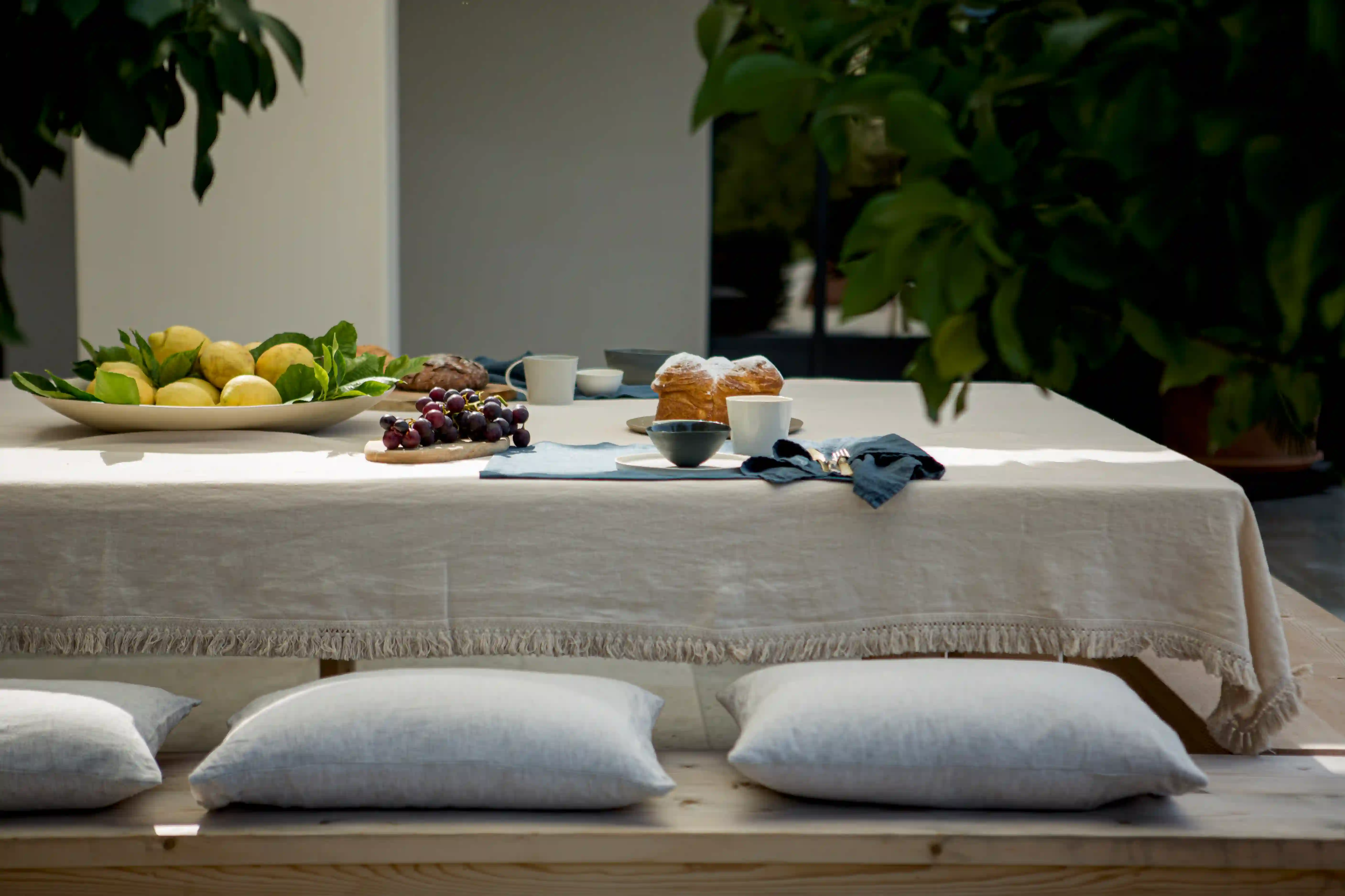 An elegant outdoor dining setup with a white fringed linen tablecloth by Once Milano, surrounded by soft linen cushions and topped with fresh fruit and pastries.