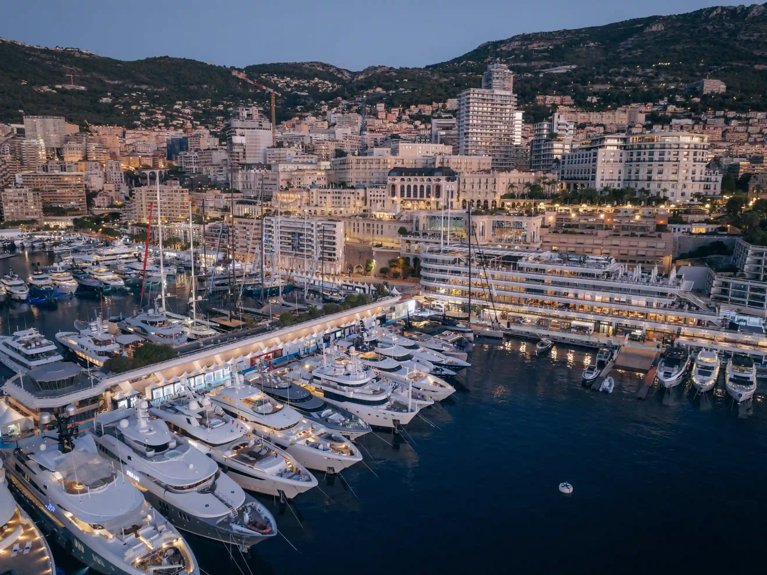 An aerial evening view of Port Hercule illuminated by yacht lights during the 2025 Monaco Yacht Show, with luxury vessels docked along the quay and the city of Monaco rising into the hills beyond.