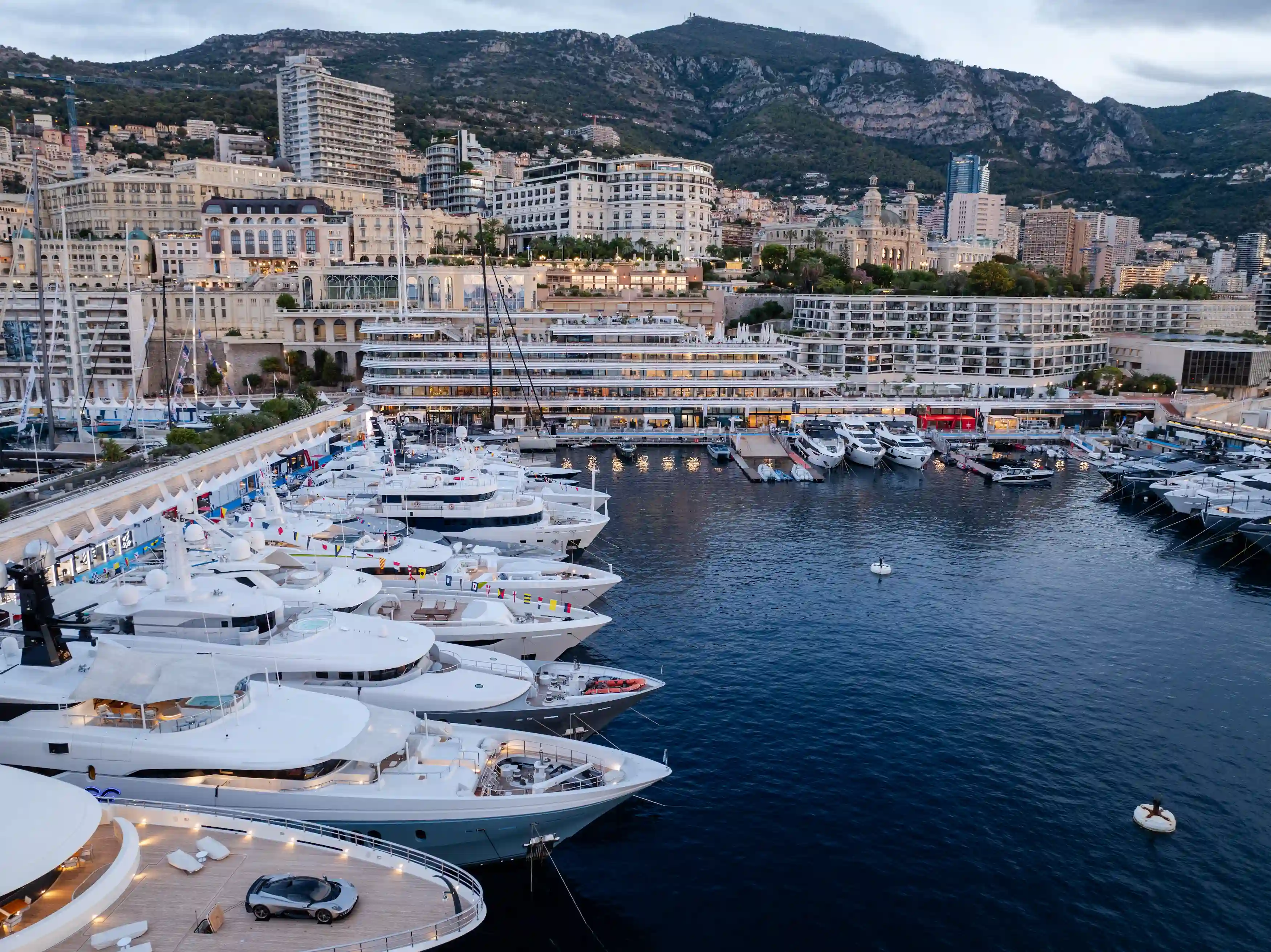 A view of luxury superyachts moored along Port Hercule during the Monaco Yacht Show 2025, with Monte Carlo’s iconic buildings and hills in the background at dusk.