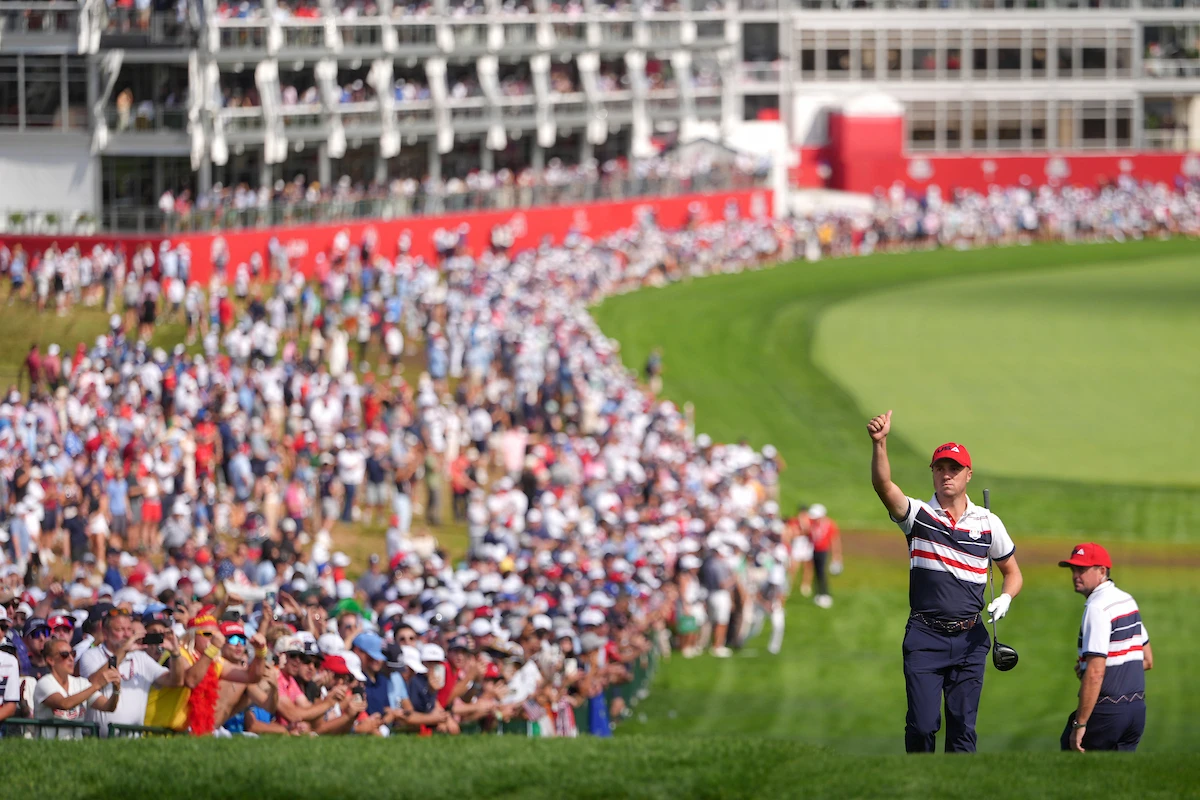 A Team USA golfer gives a thumbs-up to the crowd during the 2025 Ryder Cup at Bethpage Black, with thousands of spectators lining the fairway and grandstands in the background.