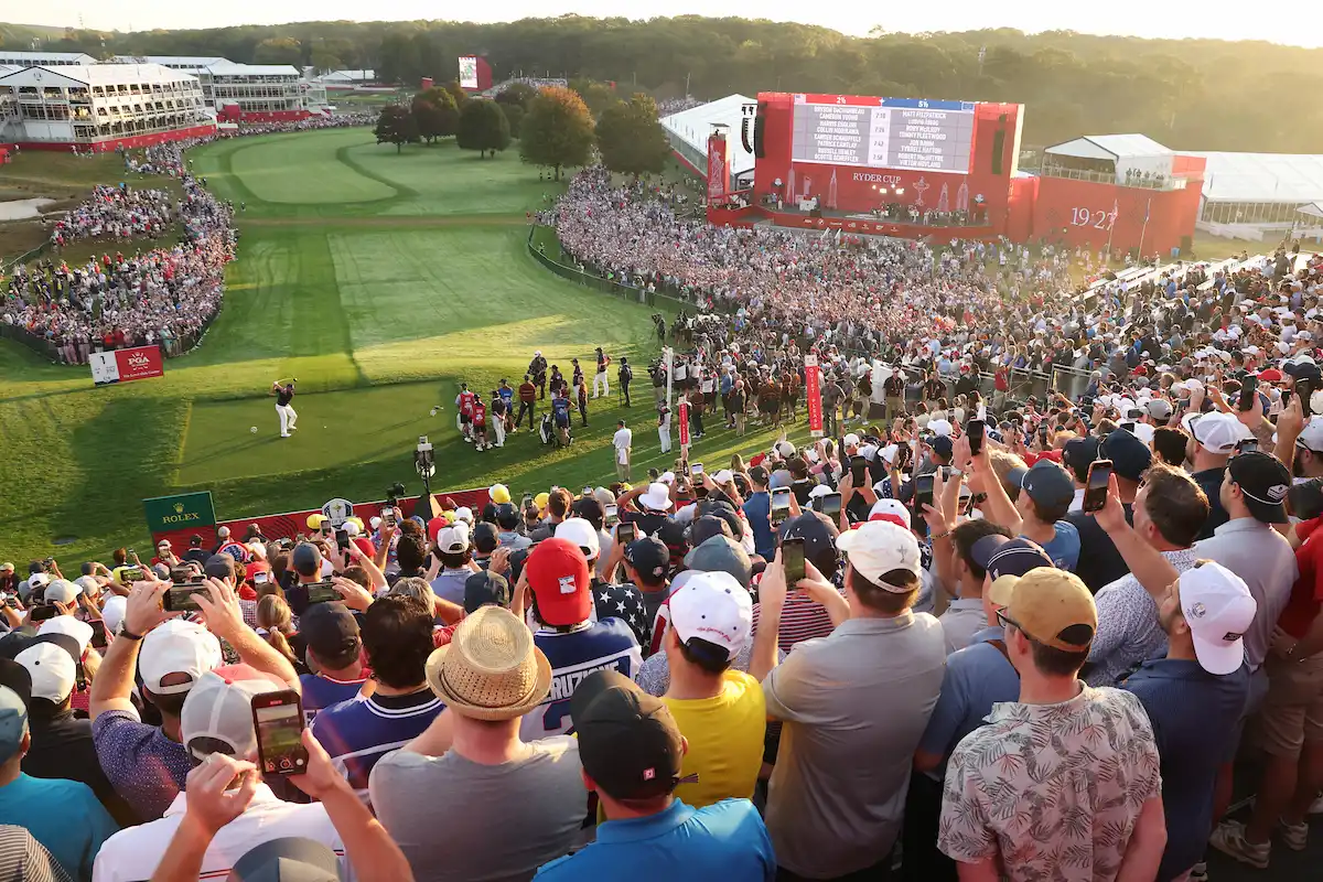Thousands of fans gather around the 1st tee at Bethpage Black during the 2025 Ryder Cup, capturing the moment on their phones as a Team USA player tees off amid roaring cheers and a vibrant crowd.