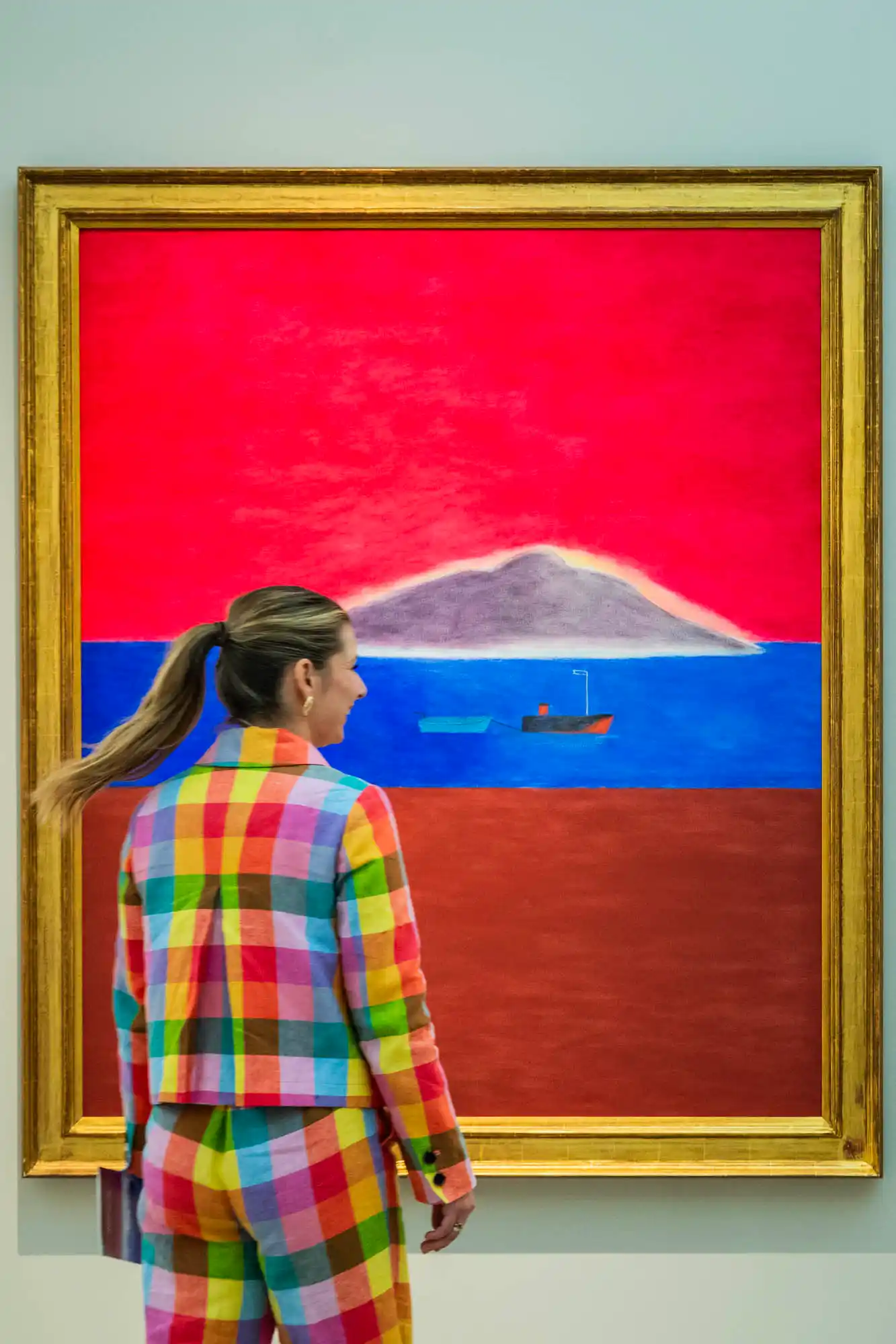 A woman in a colorful checkered suit stands in front of a vibrant seascape painting with a red sky, blue sea, and distant island at the British Art Fair 2025, capturing the energy and creativity of modern British art.