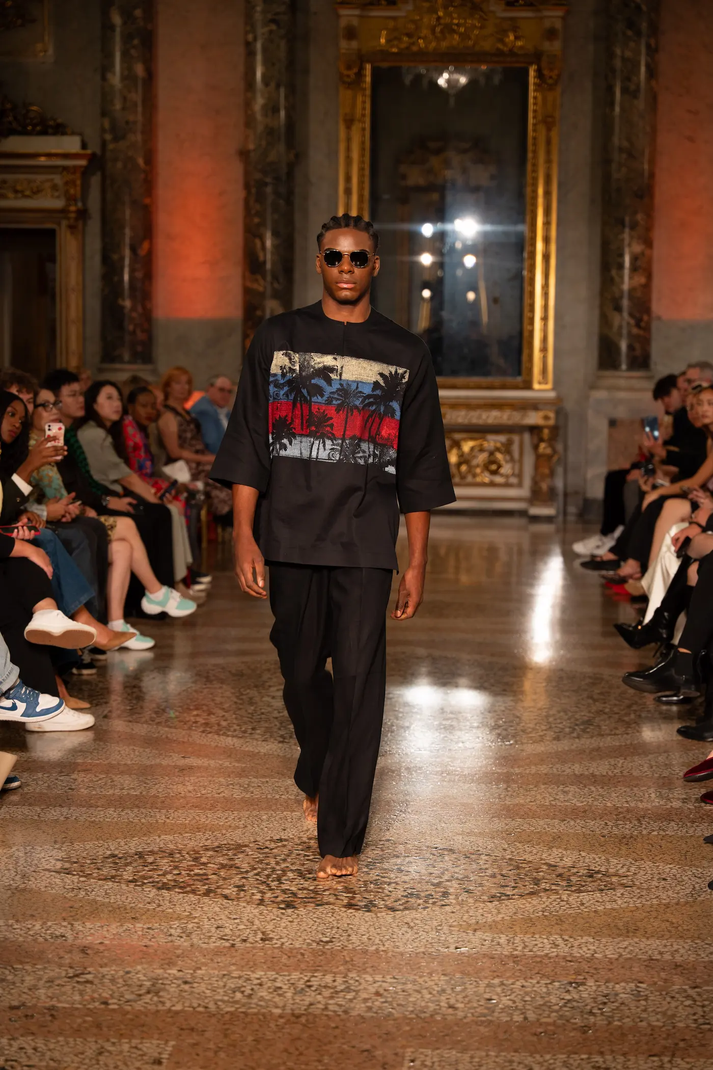 Male model walking barefoot in a black outfit with a palm tree landscape print, part of The Traveller collection.
