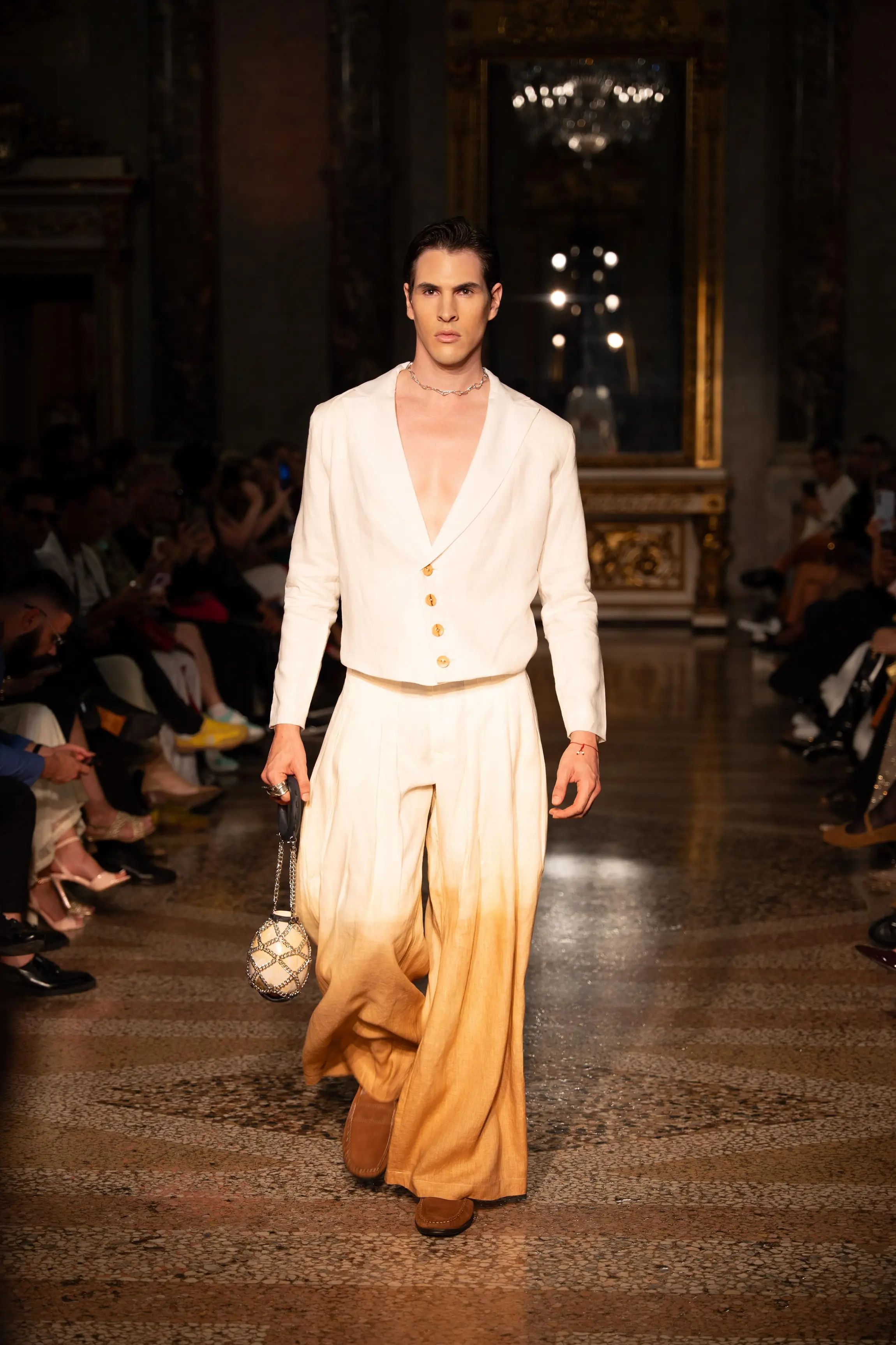 A male model walks the runway wearing a cream-to-amber gradient suit with a deep-cut blazer and wide trousers, accessorized with a beaded handbag during a high-fashion show in Milan.