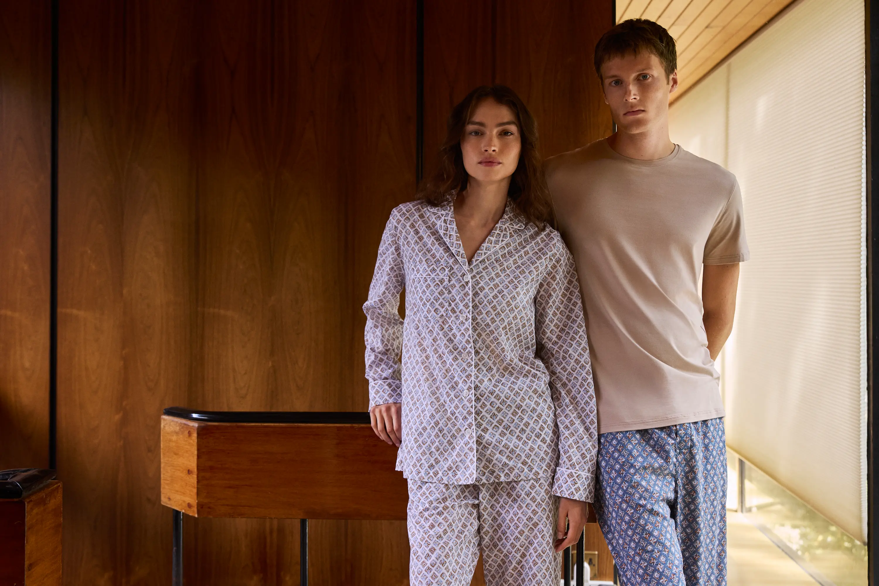 A woman in patterned cotton pyjamas and a man in a beige T-shirt and blue printed trousers stand together in a wooden-panelled modern home, reflecting relaxed sophistication.
