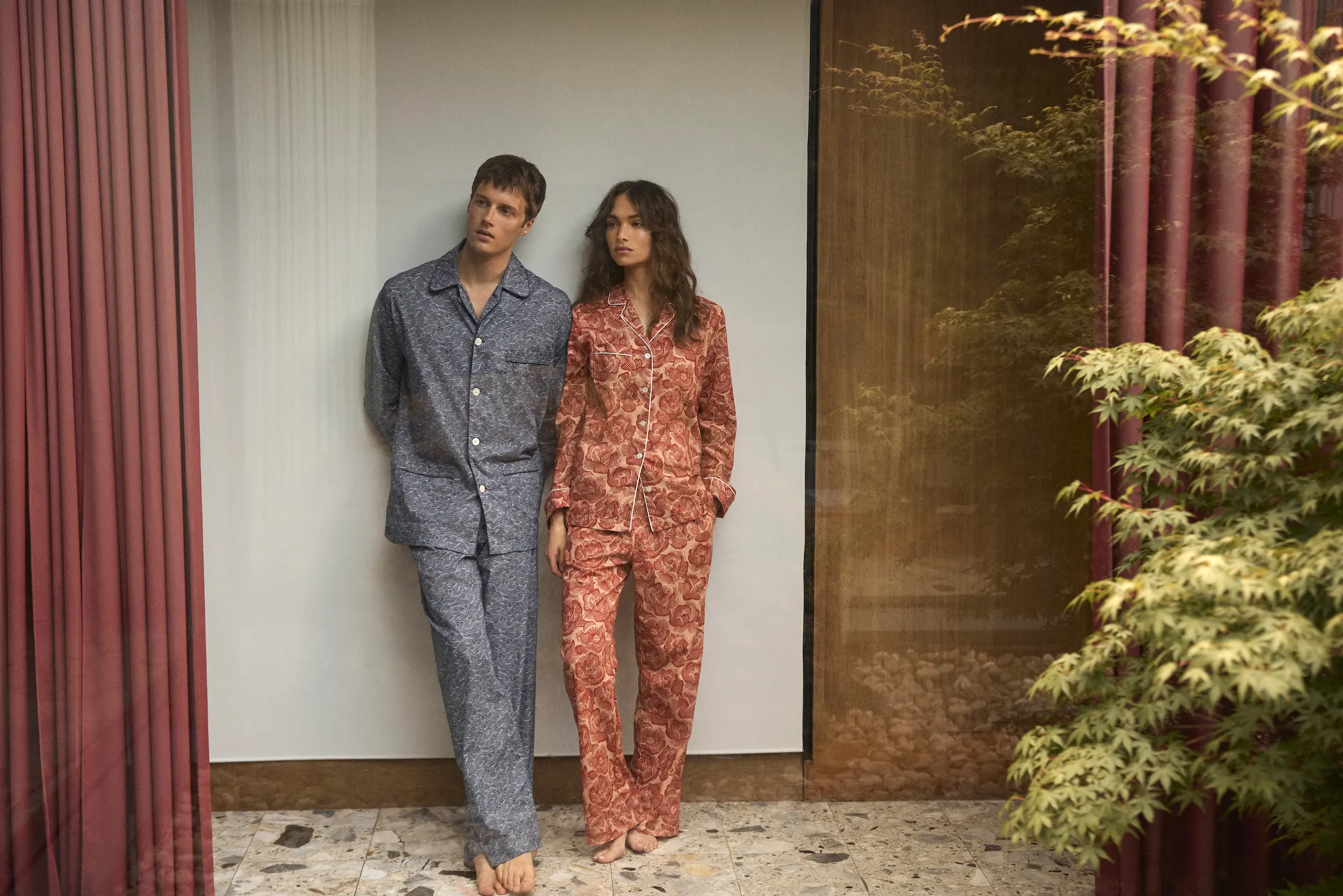 A man and woman stand together by a glass wall, wearing contrasting printed pyjamas in soft blue and coral tones from Derek Rose’s winter collection.