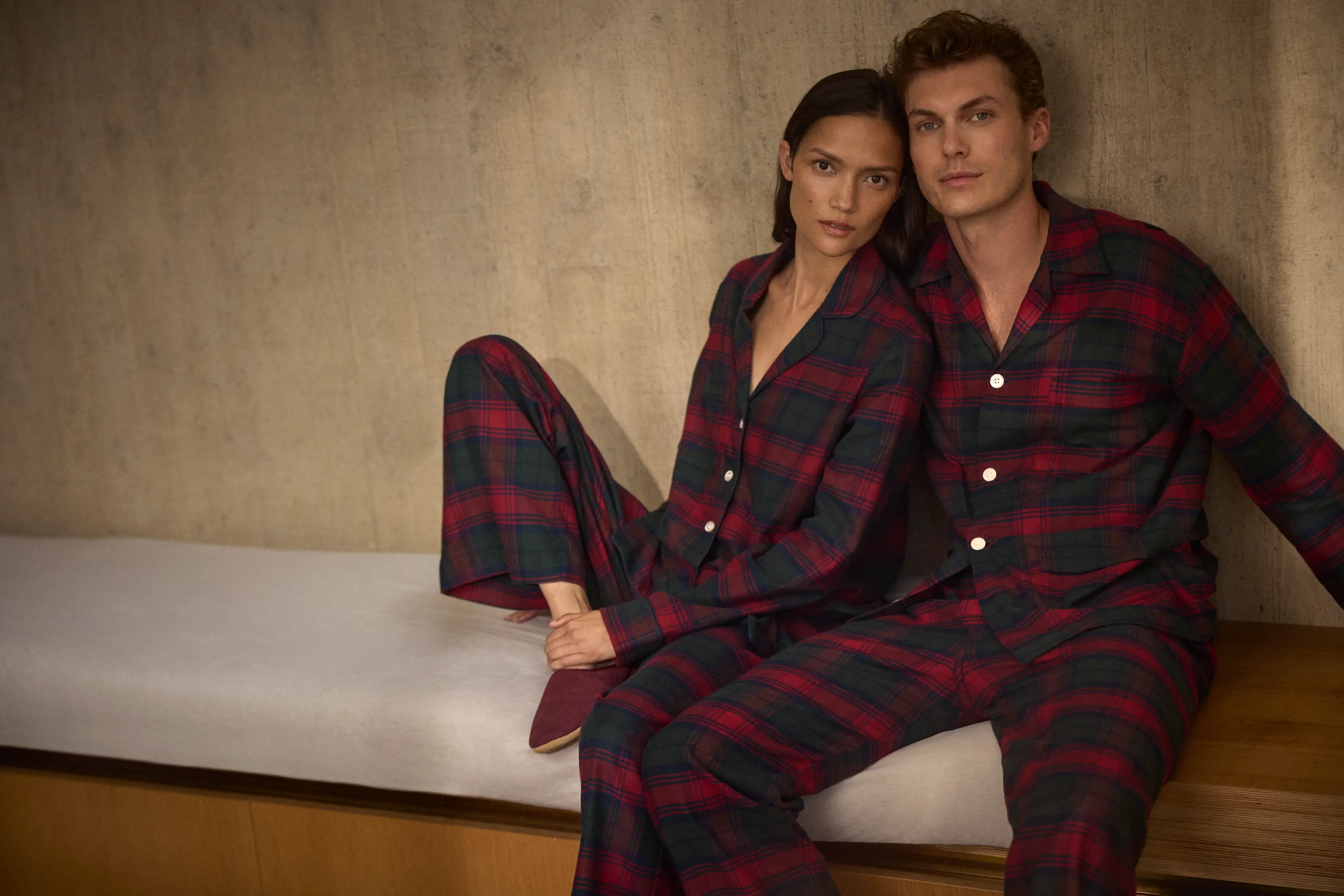 A couple wearing matching red and green tartan cotton pyjamas by Derek Rose sit together in a warm, modern interior, capturing the timeless charm of winter comfort.