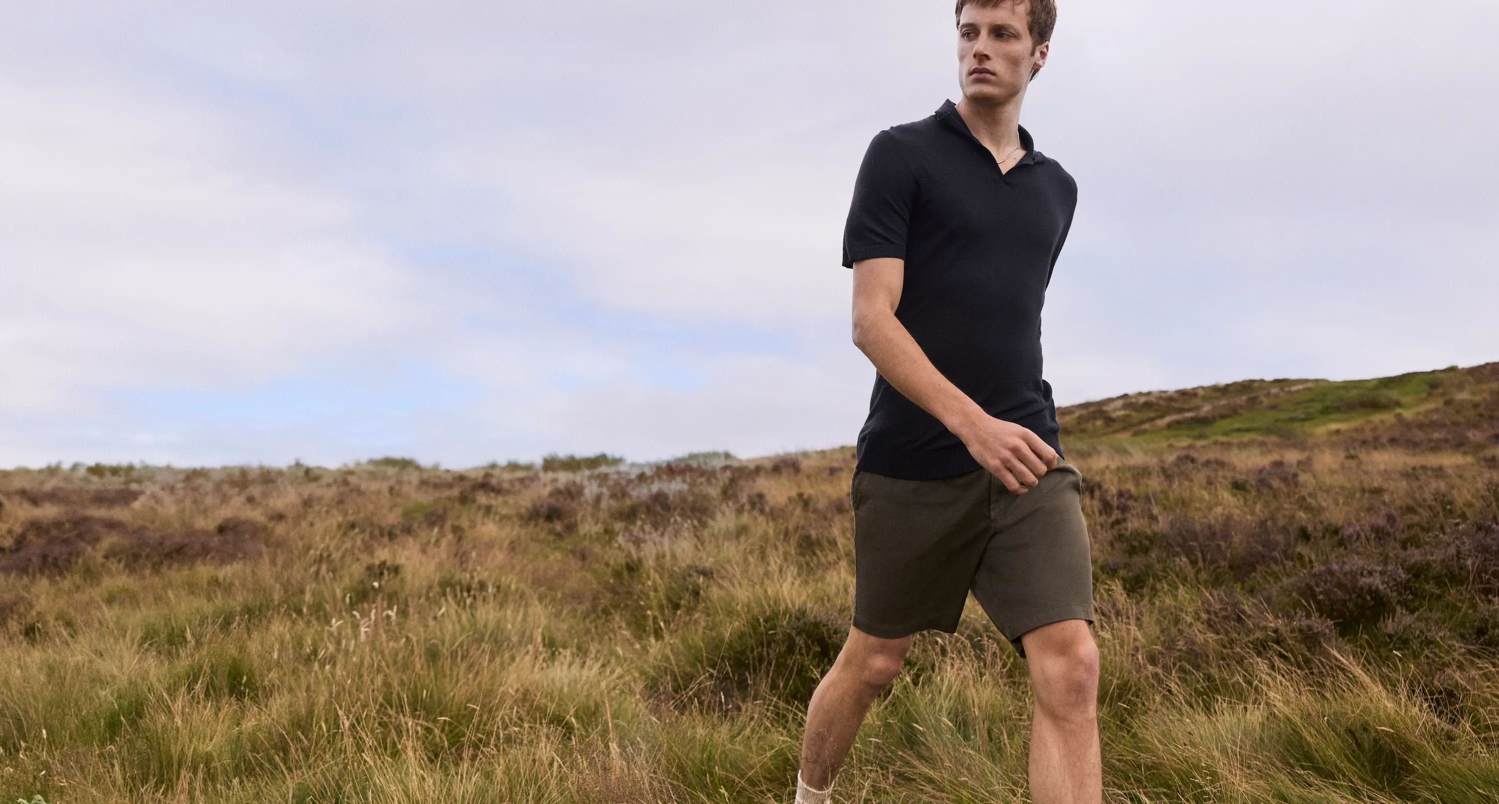 A man walks through a sunlit field wearing an elegant black merino polo shirt and olive shorts, capturing the minimalist sophistication of Derek Rose daywear.