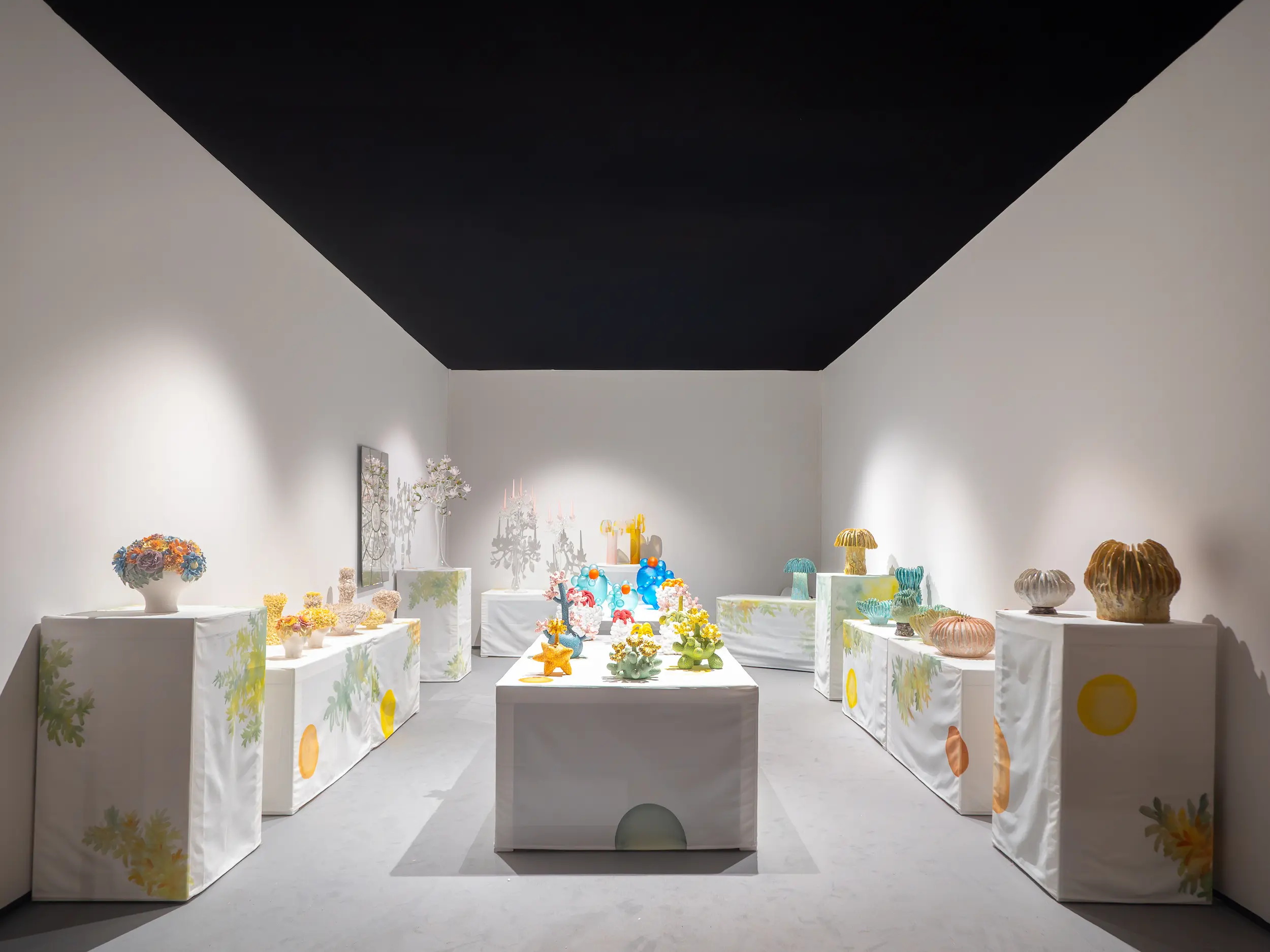 A brightly lit exhibition booth by PIK’D Gallery at PAD London 2025 featuring colorful glass and ceramic artworks arranged on white pedestals covered with hand-painted fabrics, showcasing contemporary craftsmanship and artistic vibrancy.