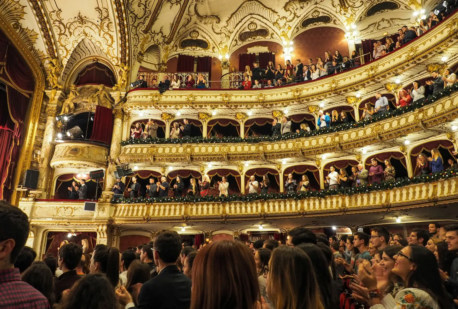Audience members give a standing ovation inside a grand, ornate theatre with red velvet curtains, golden balconies, and intricate baroque architecture.