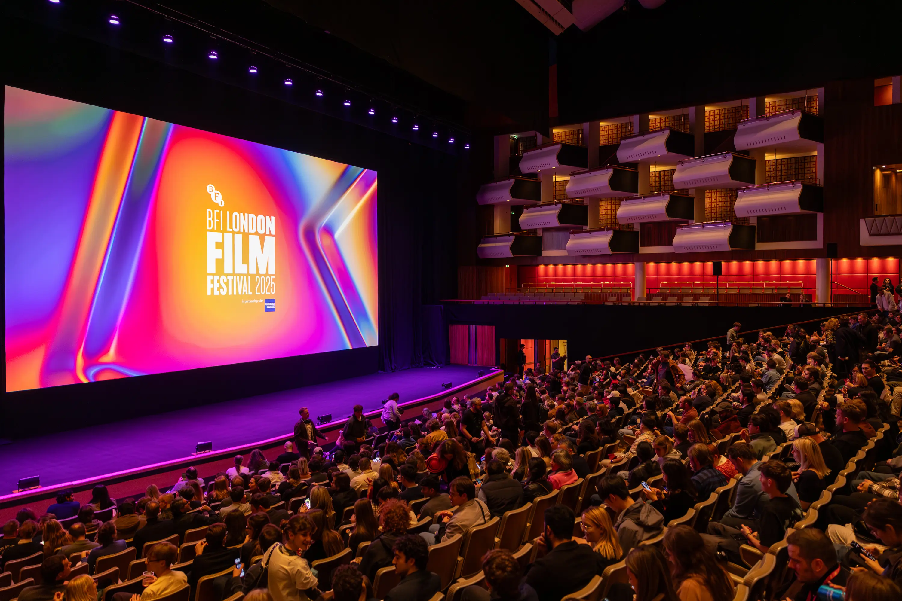 A packed audience inside the Southbank Centre’s Royal Festival Hall during the 69th BFI London Film Festival 2025, with the vibrant festival screen glowing before the premiere.