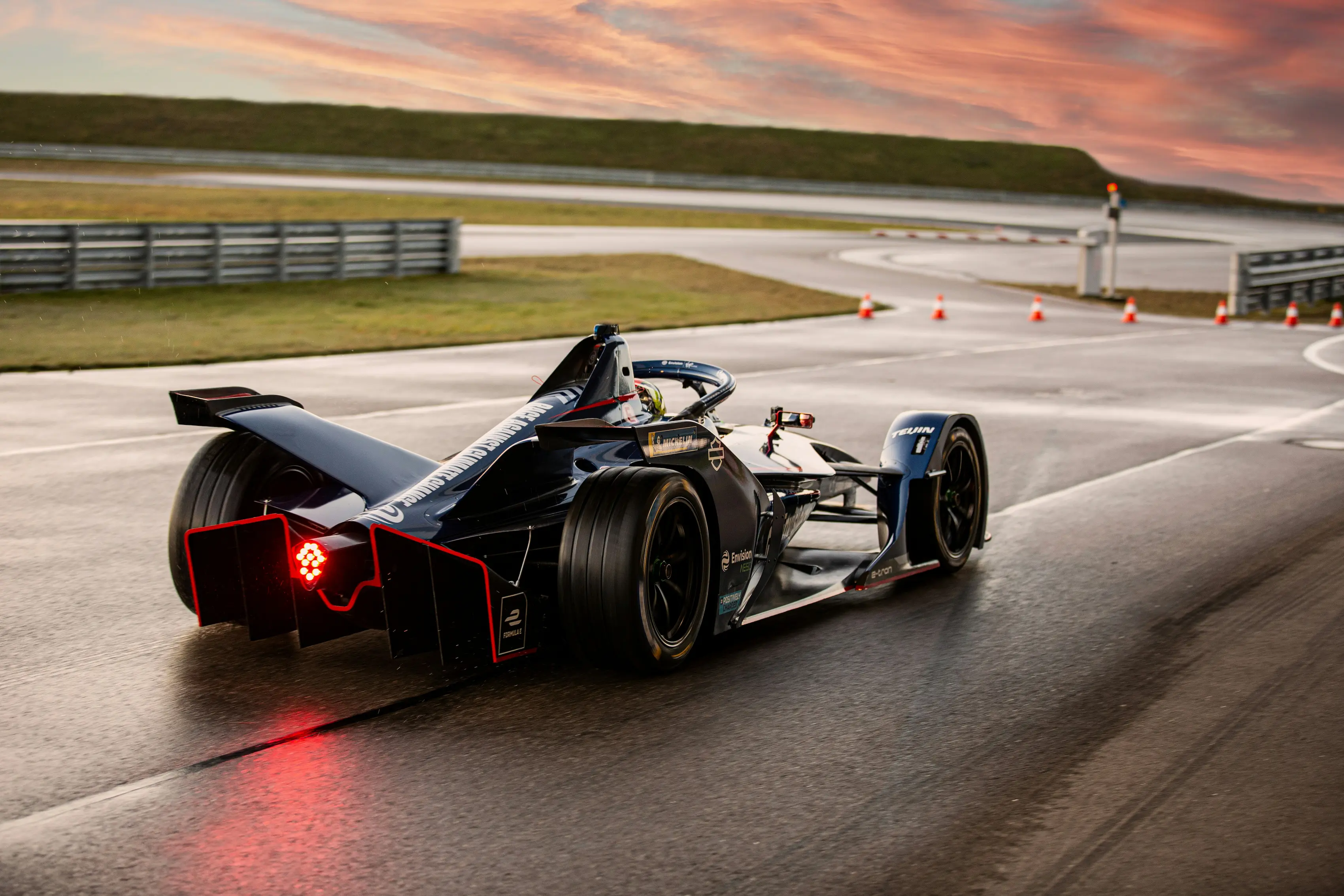 A Formula E race car speeding along a wet track during sunset, reflecting the sky’s warm colors on the asphalt.
