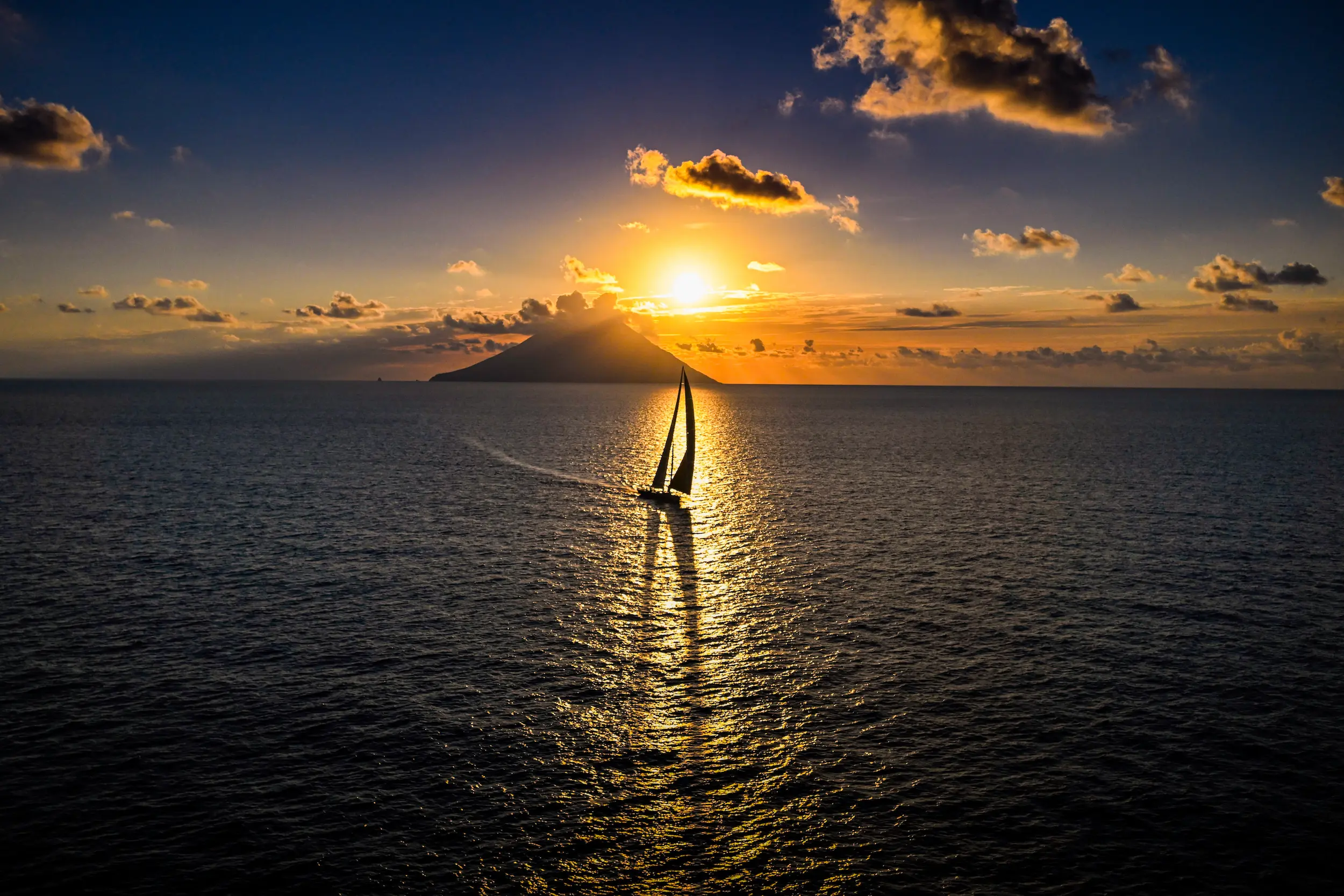 Sailing yacht silhouetted against a golden sunset near Stromboli volcano during the Rolex Middle Sea Race 2025 in the Mediterranean.