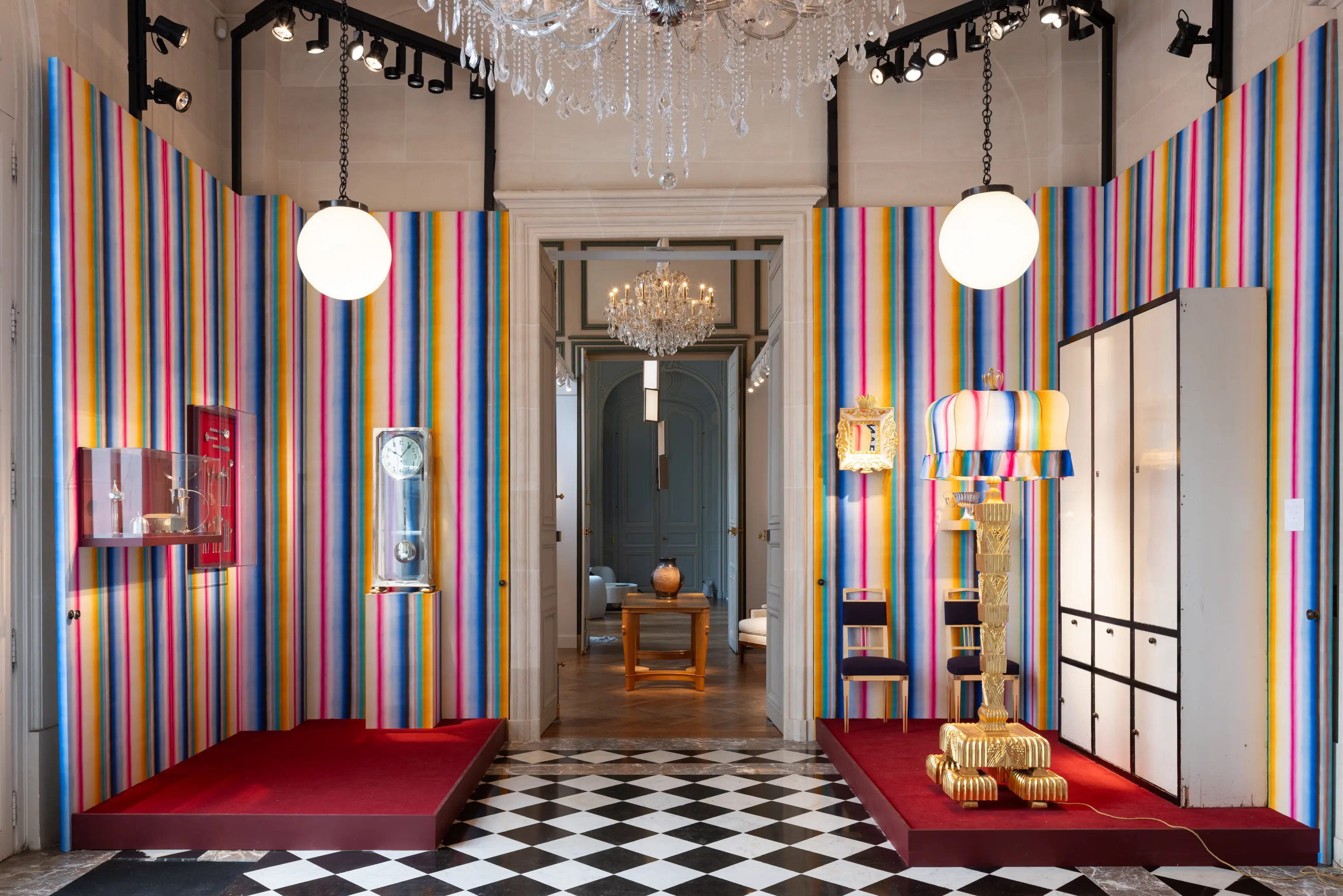 A vibrant exhibition space by Yves Macaux Gallery at Design Miami.Paris 2025, featuring colorful striped walls, Art Deco-inspired furniture, and sculptural lighting displayed under crystal chandeliers in a grand Parisian interior.
