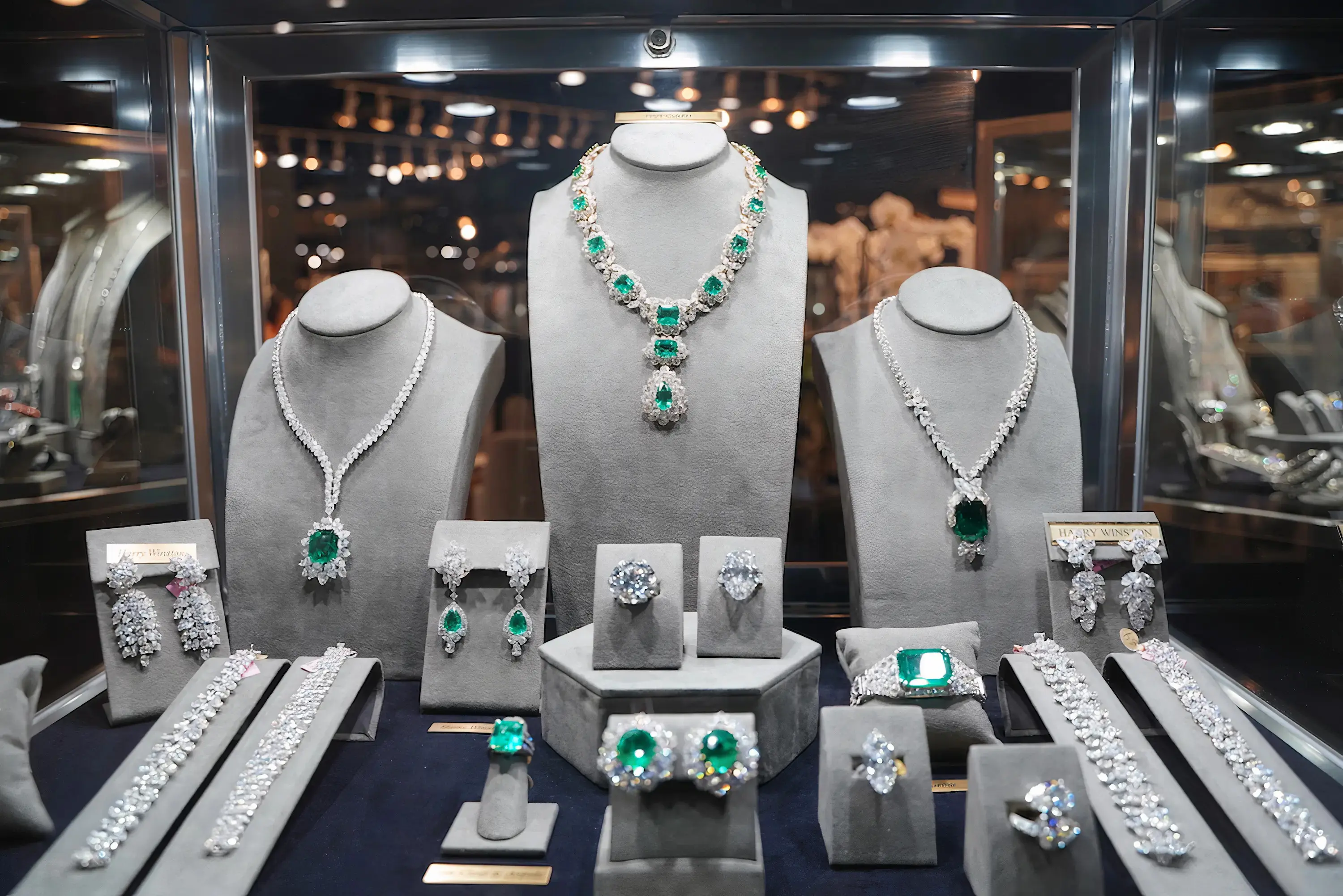 A showcase of luxury emerald and diamond jewelry, including necklaces, earrings, rings, and bracelets, displayed on grey velvet stands under elegant lighting in a high-end boutique.