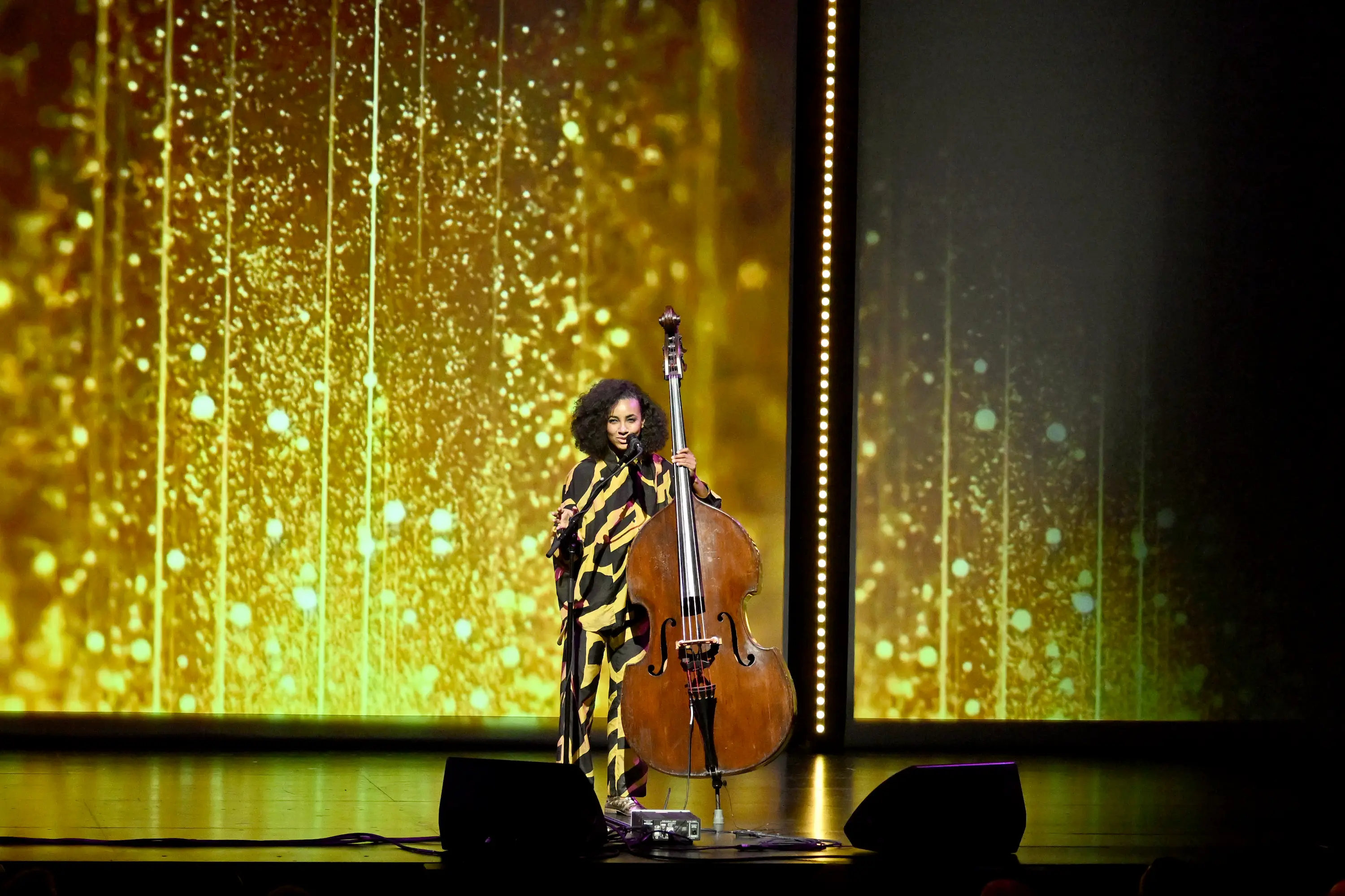 Esperanza Spalding performing on stage with her upright bass at the PAC NYC Icons of Culture Gala 2025, set against a shimmering gold backdrop.