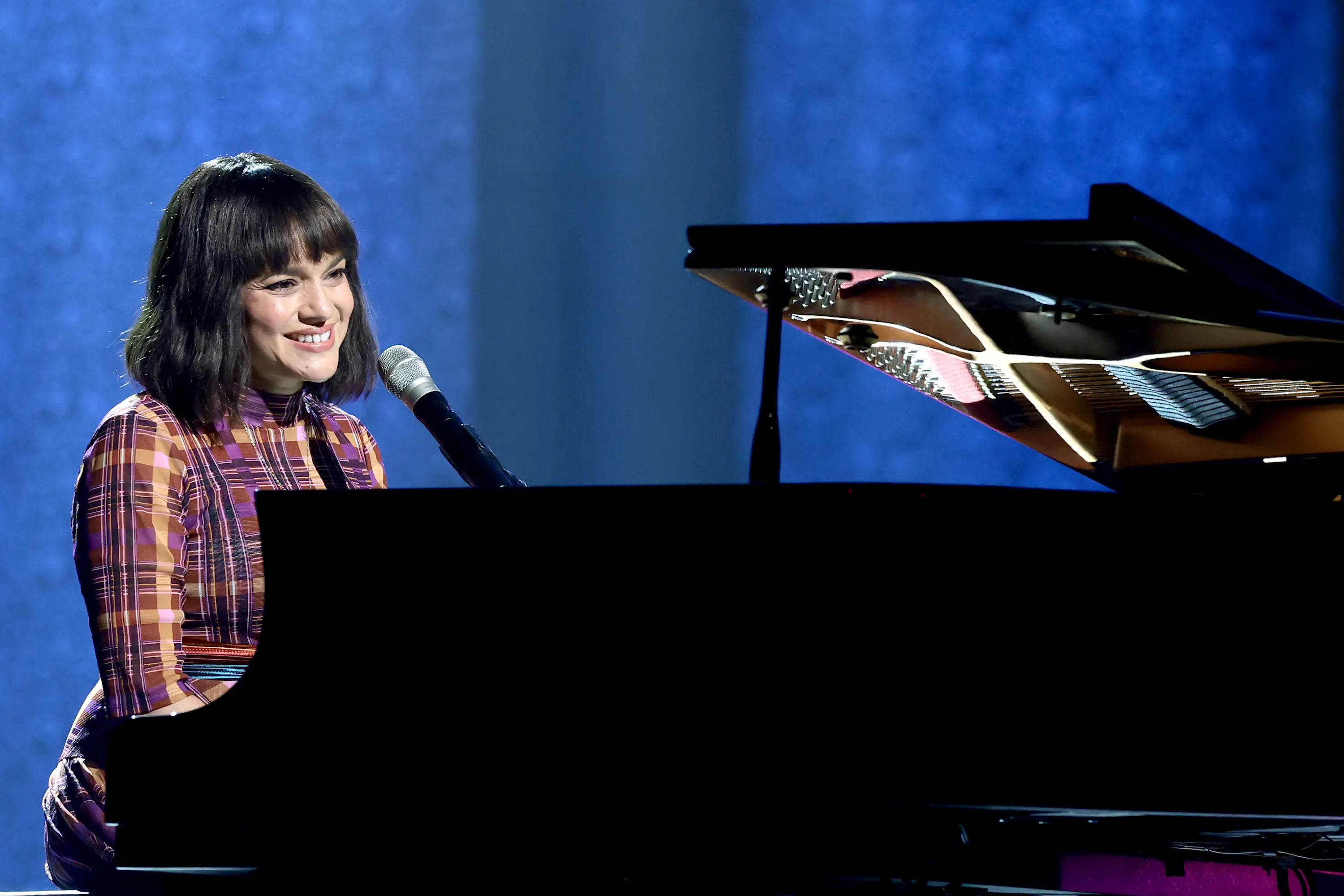 Norah Jones performing at the piano during the PAC NYC Icons of Culture Gala 2025, smiling as she sings under soft blue stage lighting.