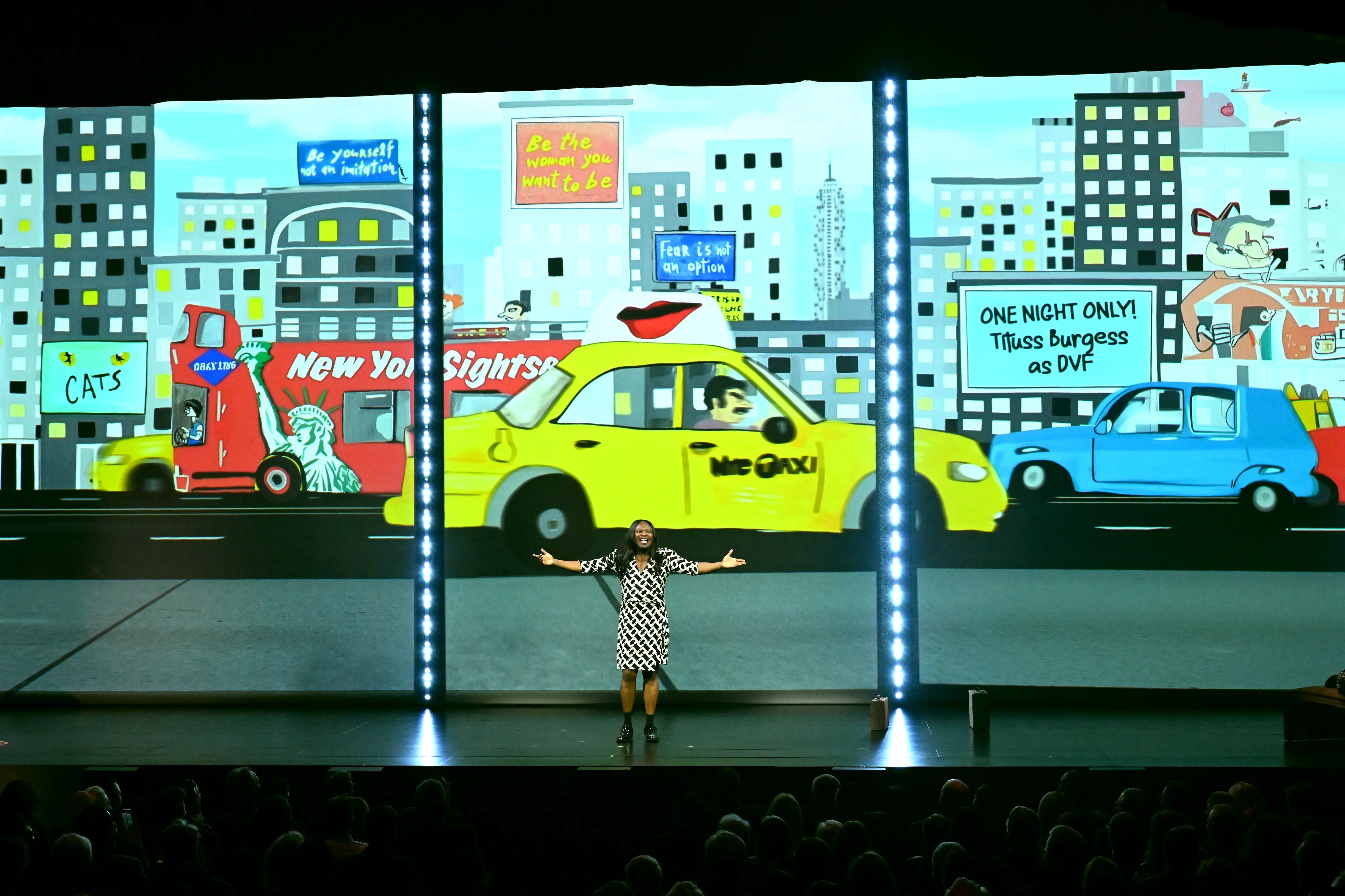 Tituss Burgess performing on stage at the PAC NYC Icons of Culture Gala 2025, standing before a colorful animated New York City backdrop featuring taxis, billboards, and city lights.