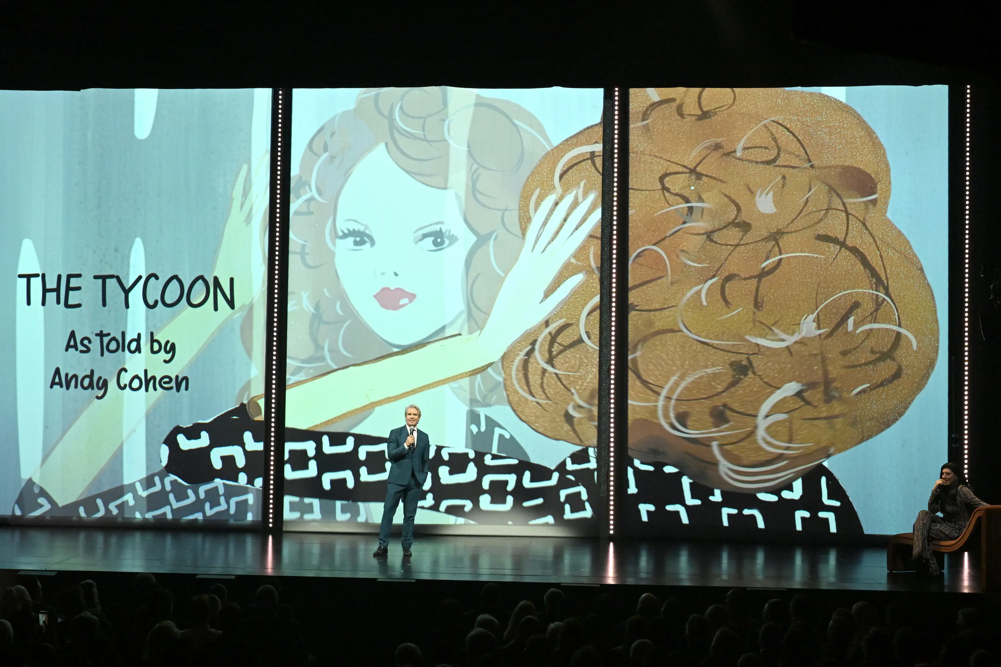 Andy Cohen speaking on stage during the PAC NYC Icons of Culture Gala 2025, standing before an illustrated backdrop reading “The Tycoon – As told by Andy Cohen,” featuring stylized artwork of a woman with voluminous hair.