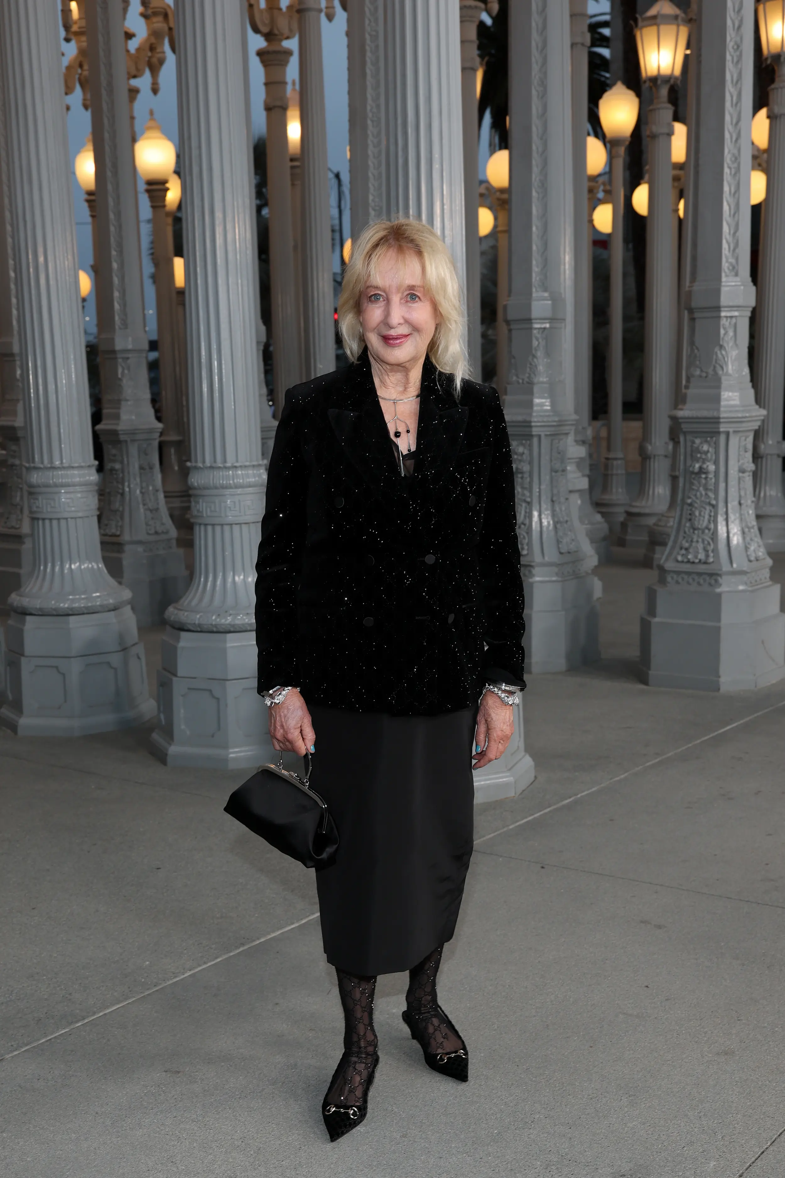 Woman in an elegant black outfit poses beneath the glowing Urban Light installation at LACMA during the Art+Film Gala evening.
