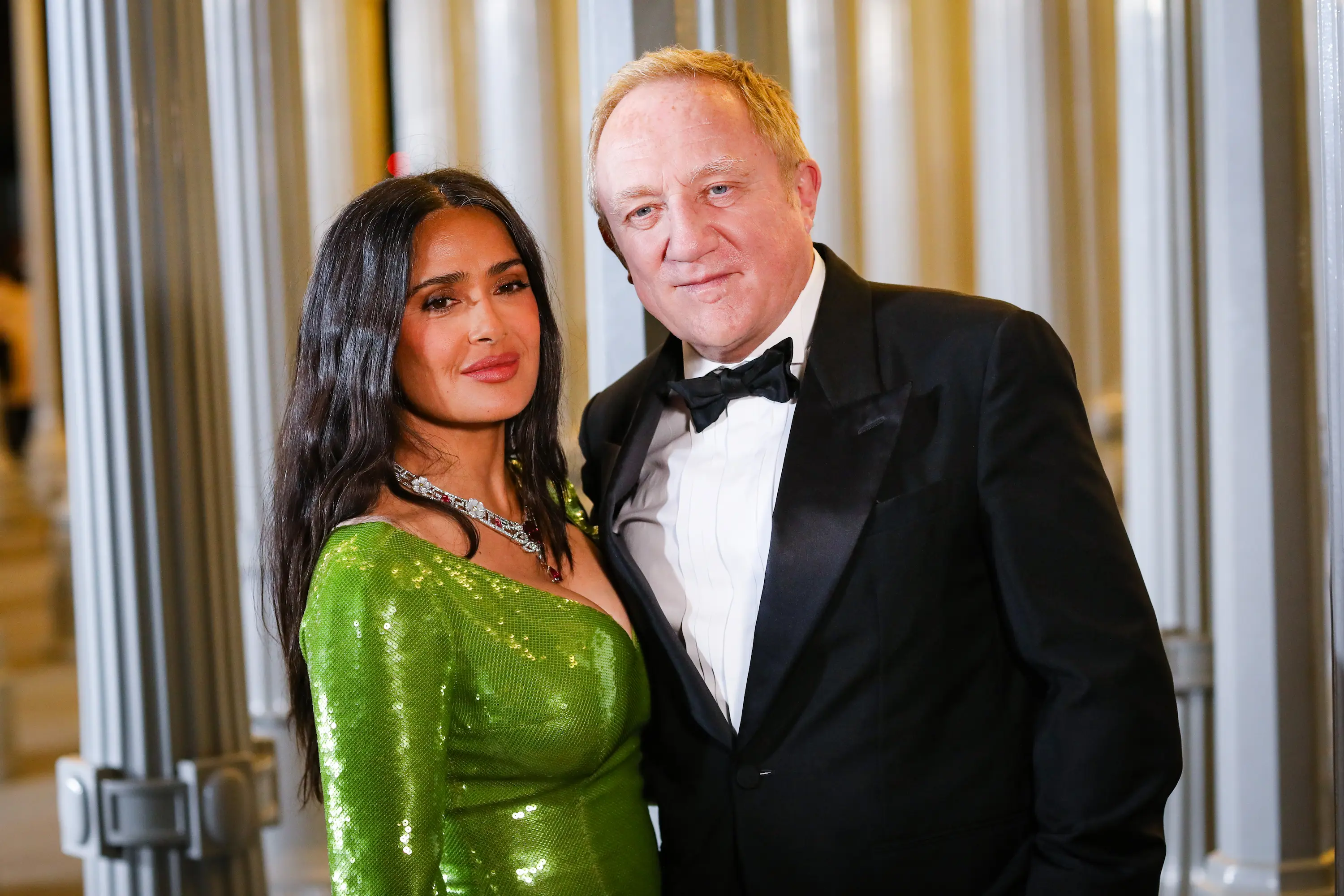 Couple poses together at the LACMA Art+Film Gala, with the woman wearing a shimmering green gown and the man in a classic black tuxedo beneath the glowing Urban Light installation.