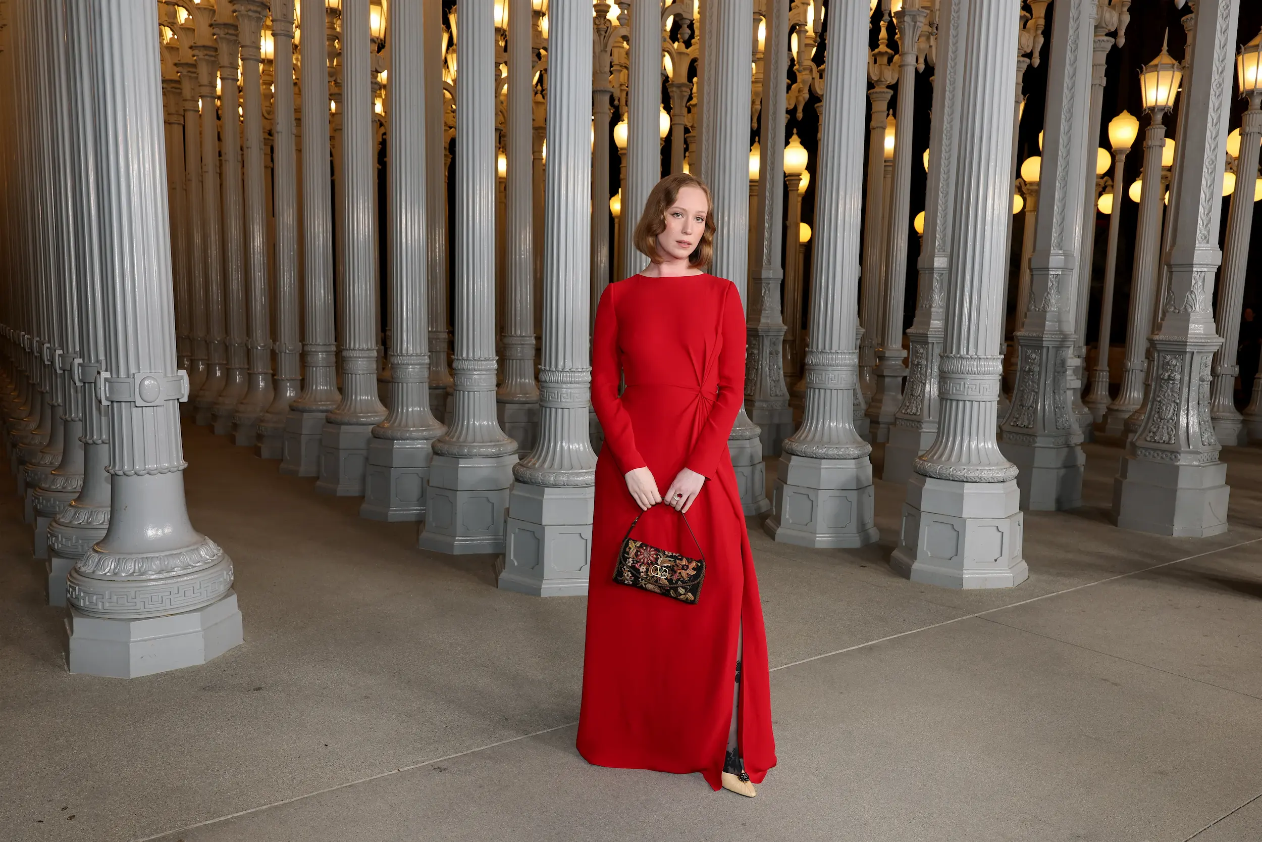 Woman wearing a long red dress and holding a floral handbag poses gracefully among the illuminated pillars of the Urban Light installation at LACMA during the Art+Film Gala.