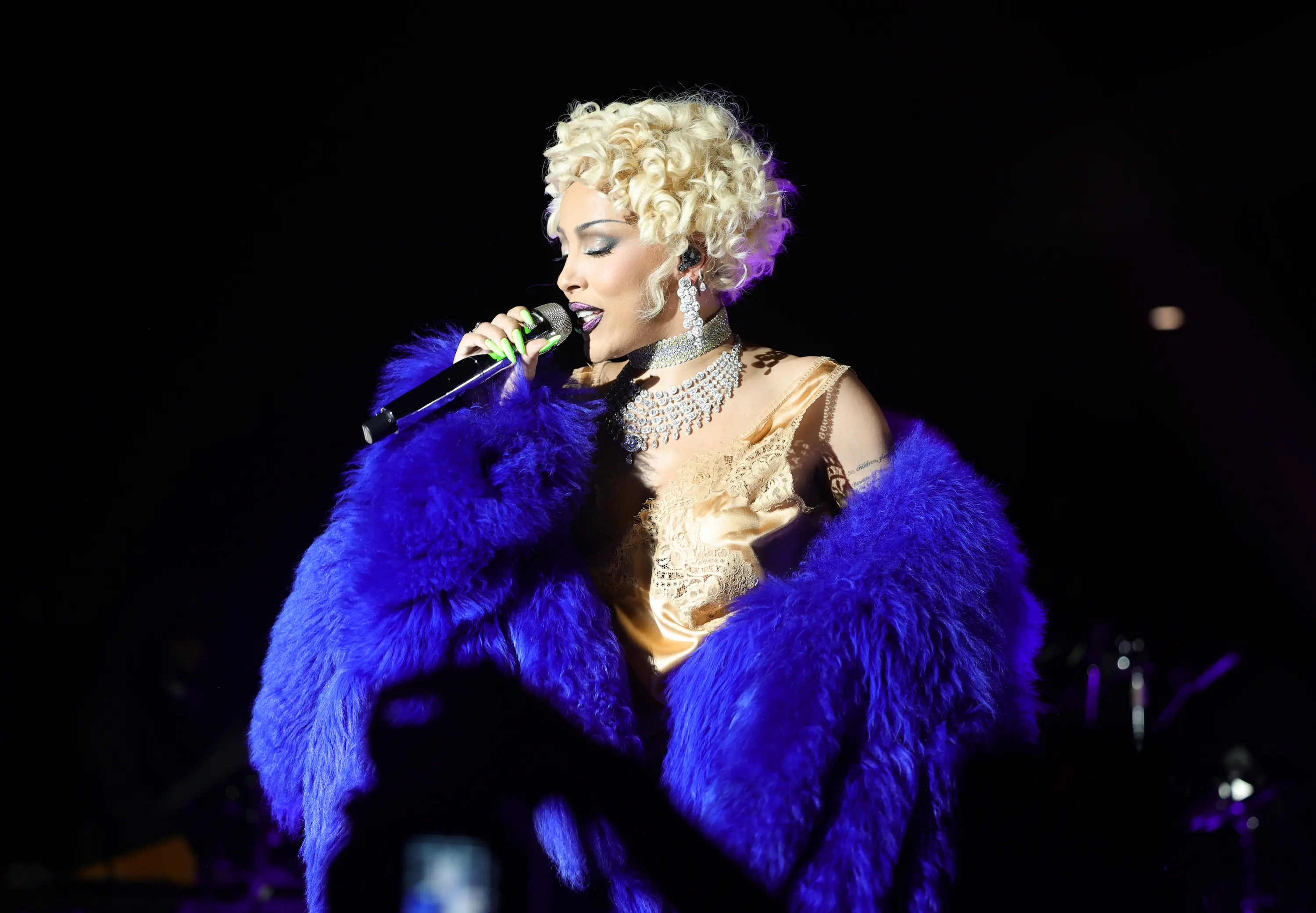 Woman with short blonde curls performs on stage wearing a dramatic royal blue fur coat, gold satin dress, and diamond jewelry during the LACMA Art+Film Gala.