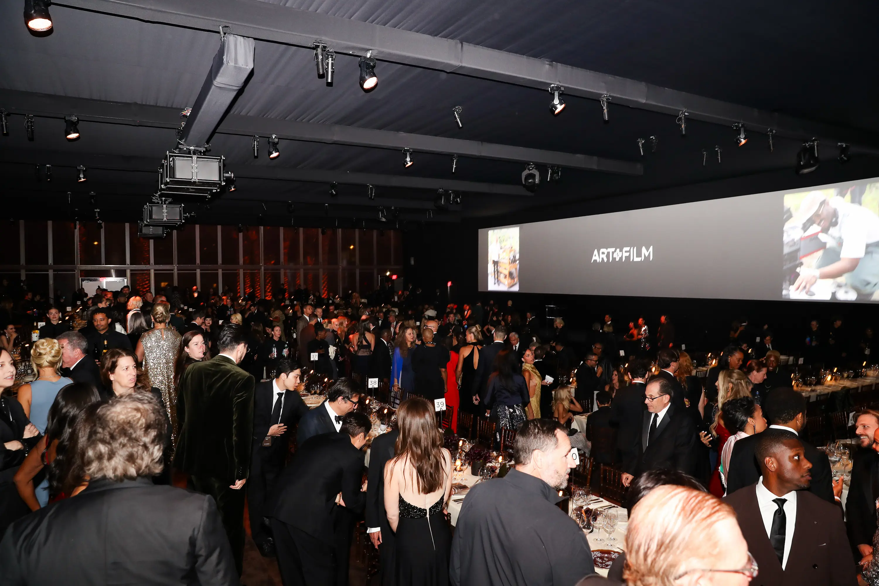 A large crowd of elegantly dressed guests mingles inside the candlelit gala tent at the LACMA Art+Film Gala, with a wide screen displaying the event’s logo in the background.