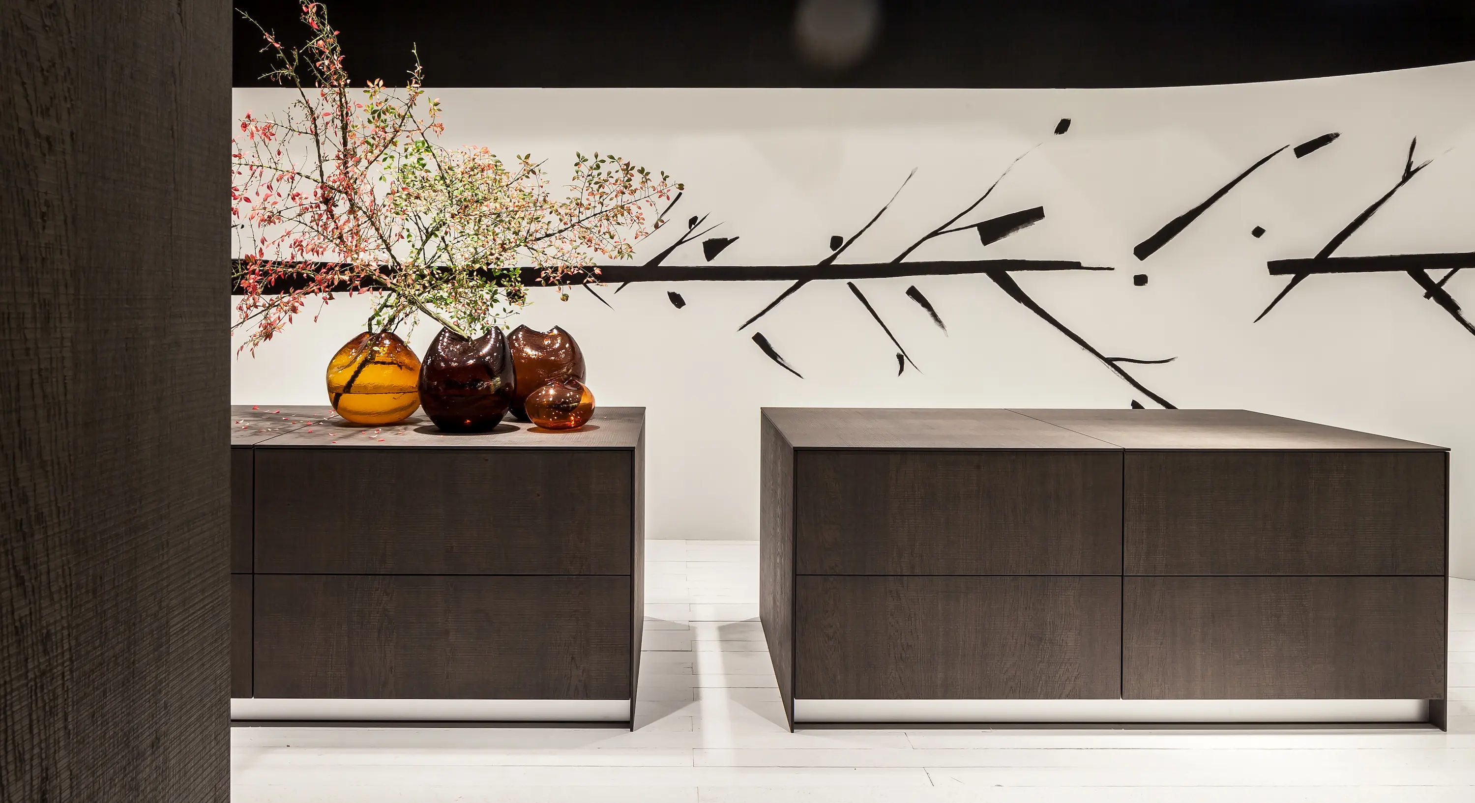 A contemporary minimalist interior featuring two dark wood cabinets adorned with amber glass vases and flowering branches, designed by When Objects Work.