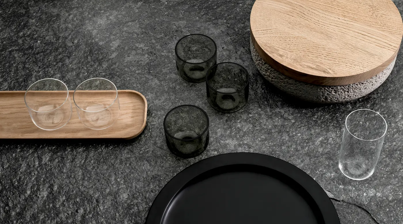 Top view of minimalist tableware by When Objects Work, including smoked glass cups, a wooden tray, a black plate, and a round stone bowl with a wooden lid on a textured dark surface.