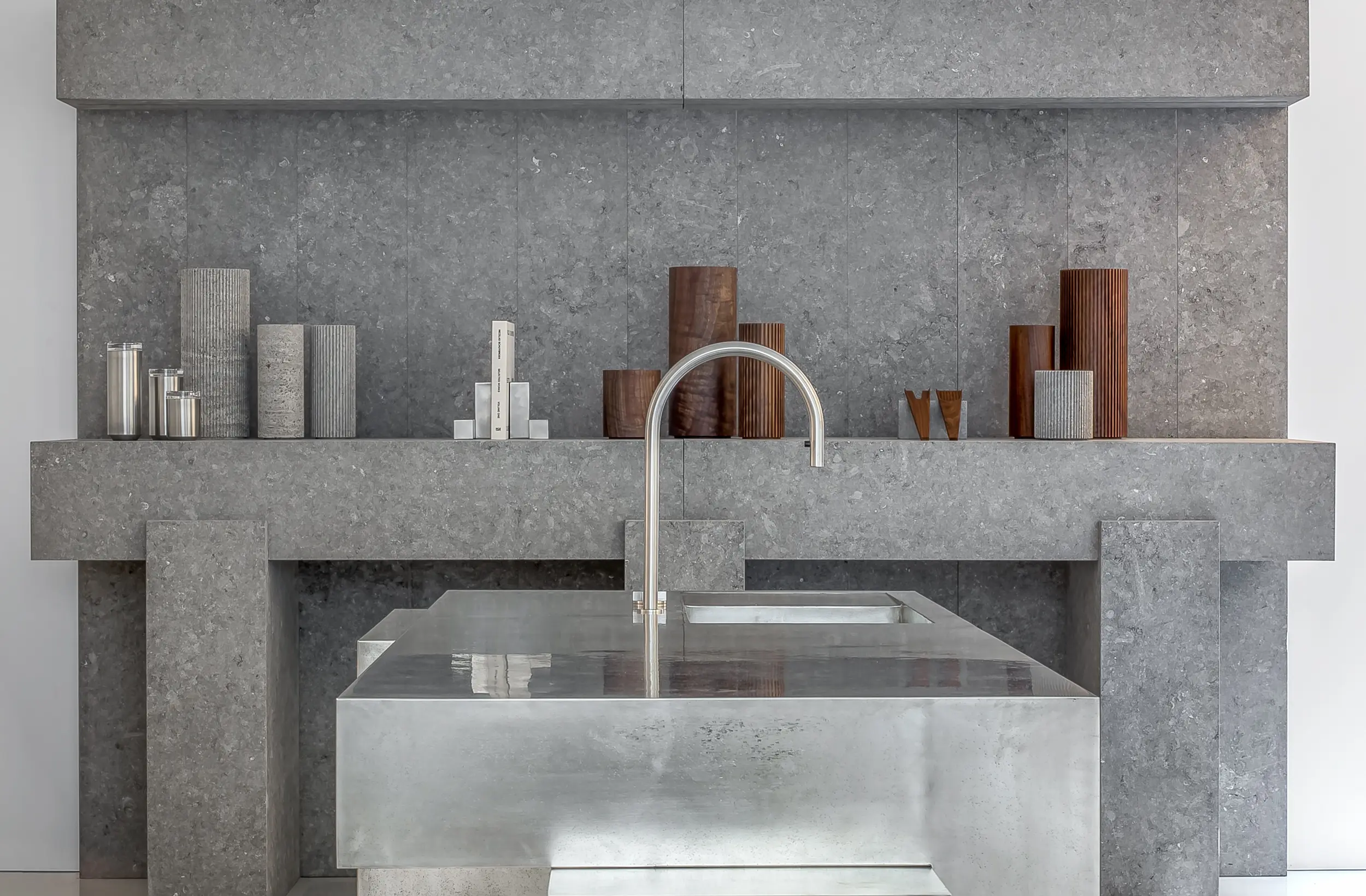 Modern kitchen interior featuring a sculptural stone wall with wooden and metal vases, a sleek stainless steel island, and minimalist faucet design by Nicholas Schuybroek for When Objects Work.