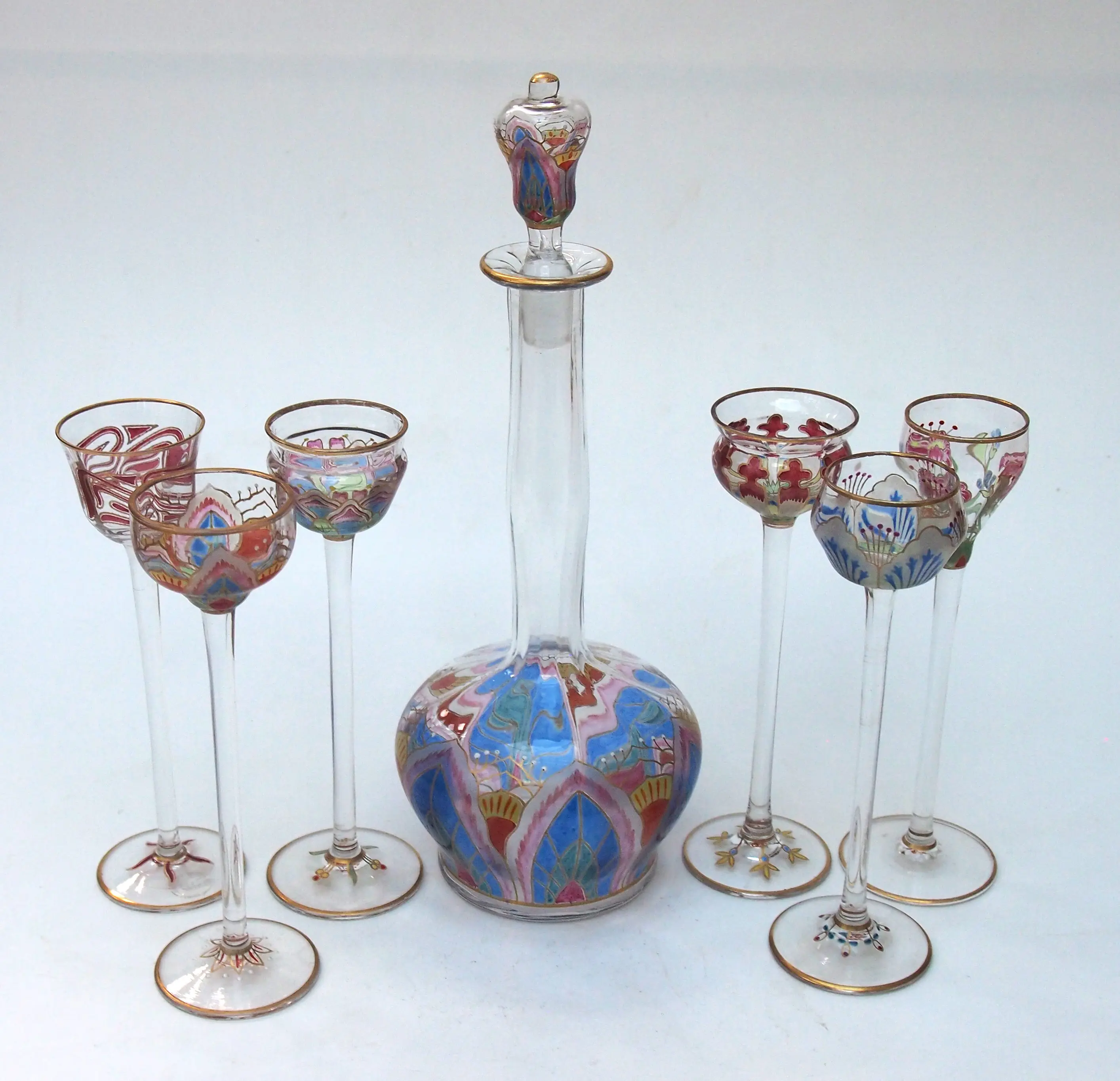 Exquisite antique liqueur set by Meyr’s Neffe, featuring a hand-painted decanter and six matching harlequin flower glasses with intricate enamel and gilded Art Nouveau motifs, presented by M&D Moir.