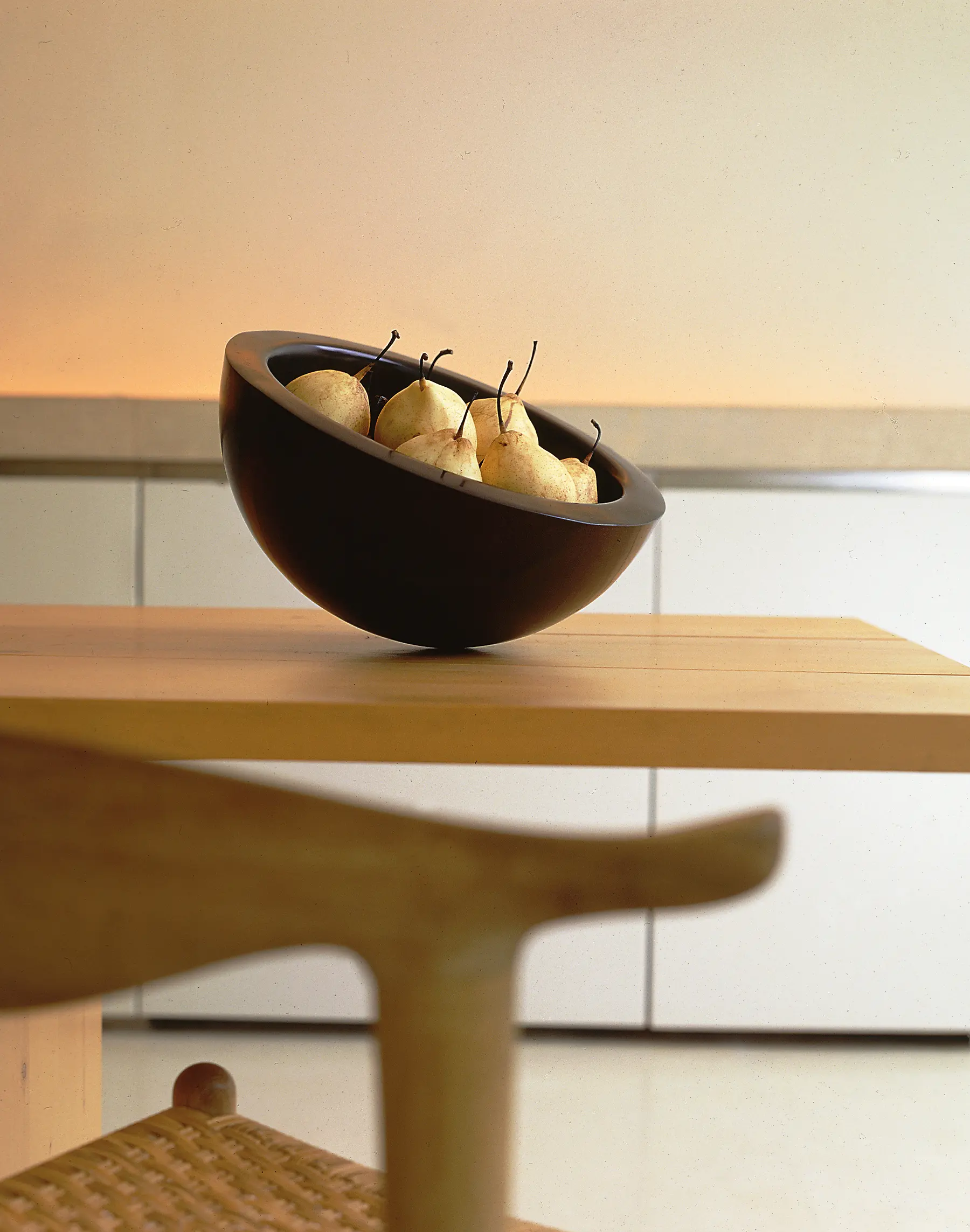 A minimalist wooden bowl filled with ripe pears sits on a light wooden table beside a woven chair in a serene modern interior.