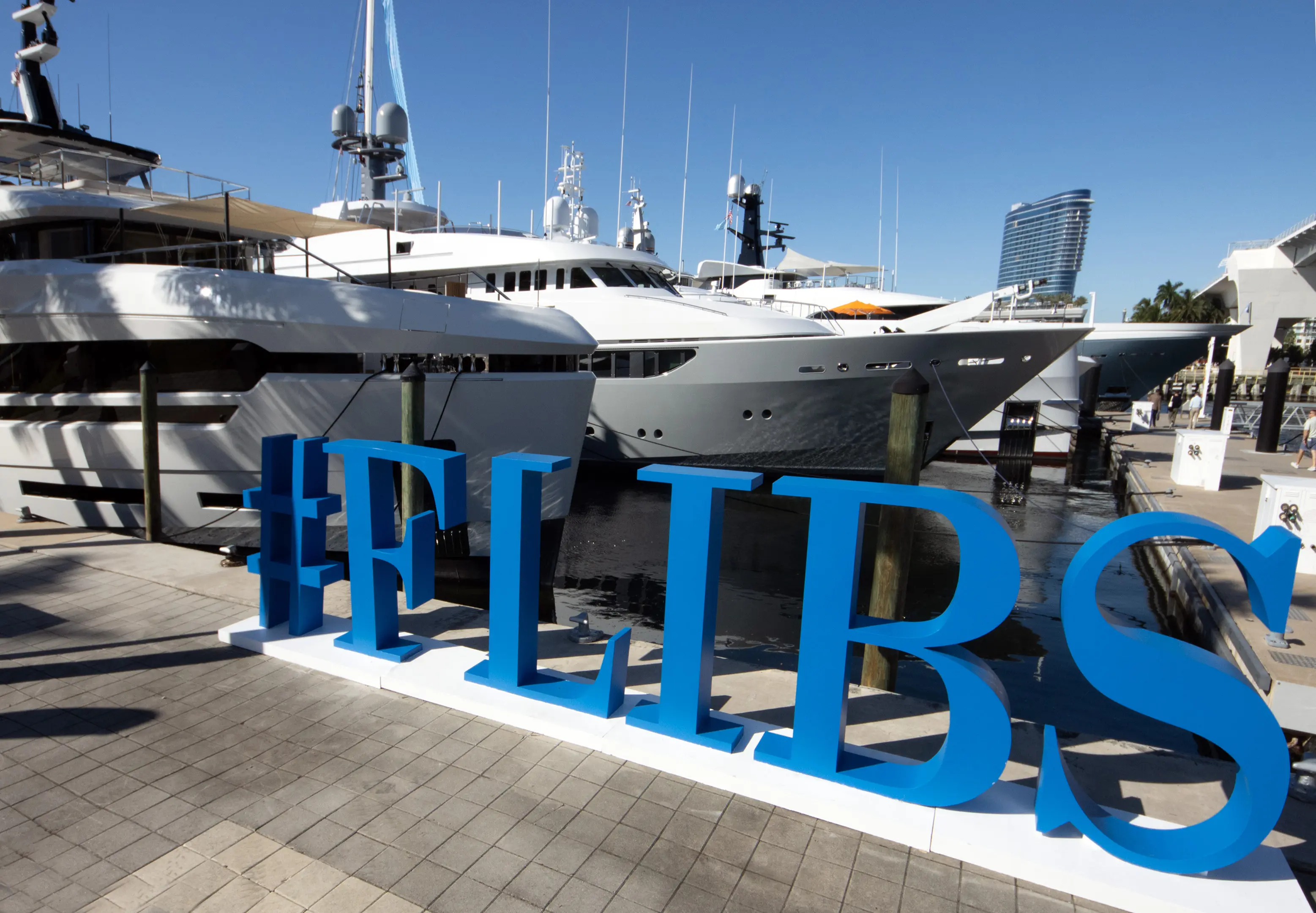 Large blue #FLIBS sign displayed in front of luxury superyachts docked at the marina during the 2025 Fort Lauderdale International Boat Show under clear skies.