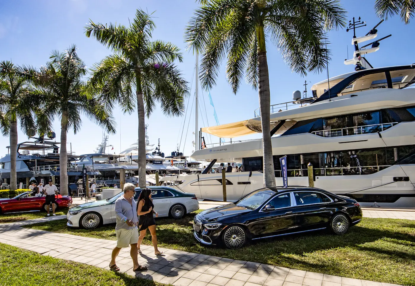 Visitors walk past luxury yachts and high-end cars displayed among palm trees at the 2025 Fort Lauderdale International Boat Show on a sunny Florida day.