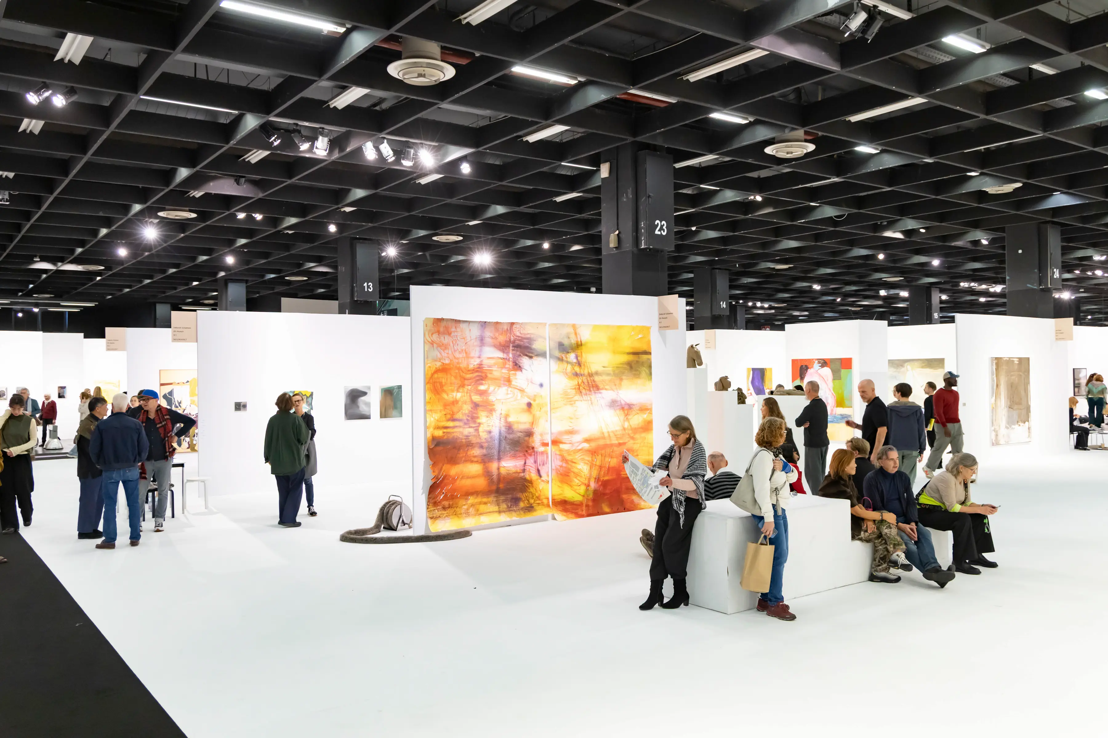 Guests walk through the exhibition halls of Art Cologne 2025, observing modern and contemporary artworks displayed in sleek white booths under the iconic Koelnmesse ceiling.