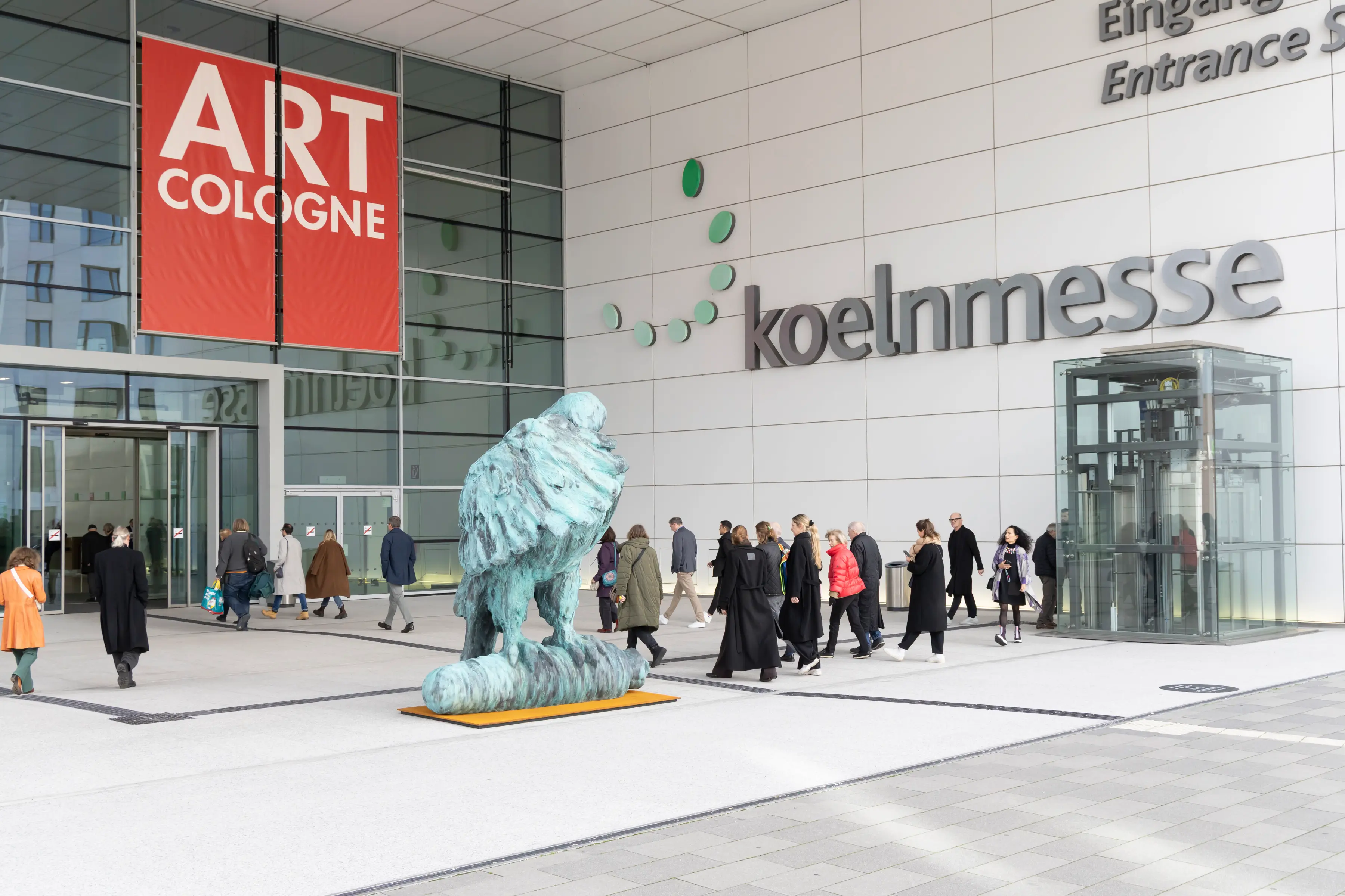Visitors enter Koelnmesse for Art Cologne 2025 beneath a large red banner, passing a monumental bronze sculpture that marks the entrance to the world’s oldest modern and contemporary art fair.
