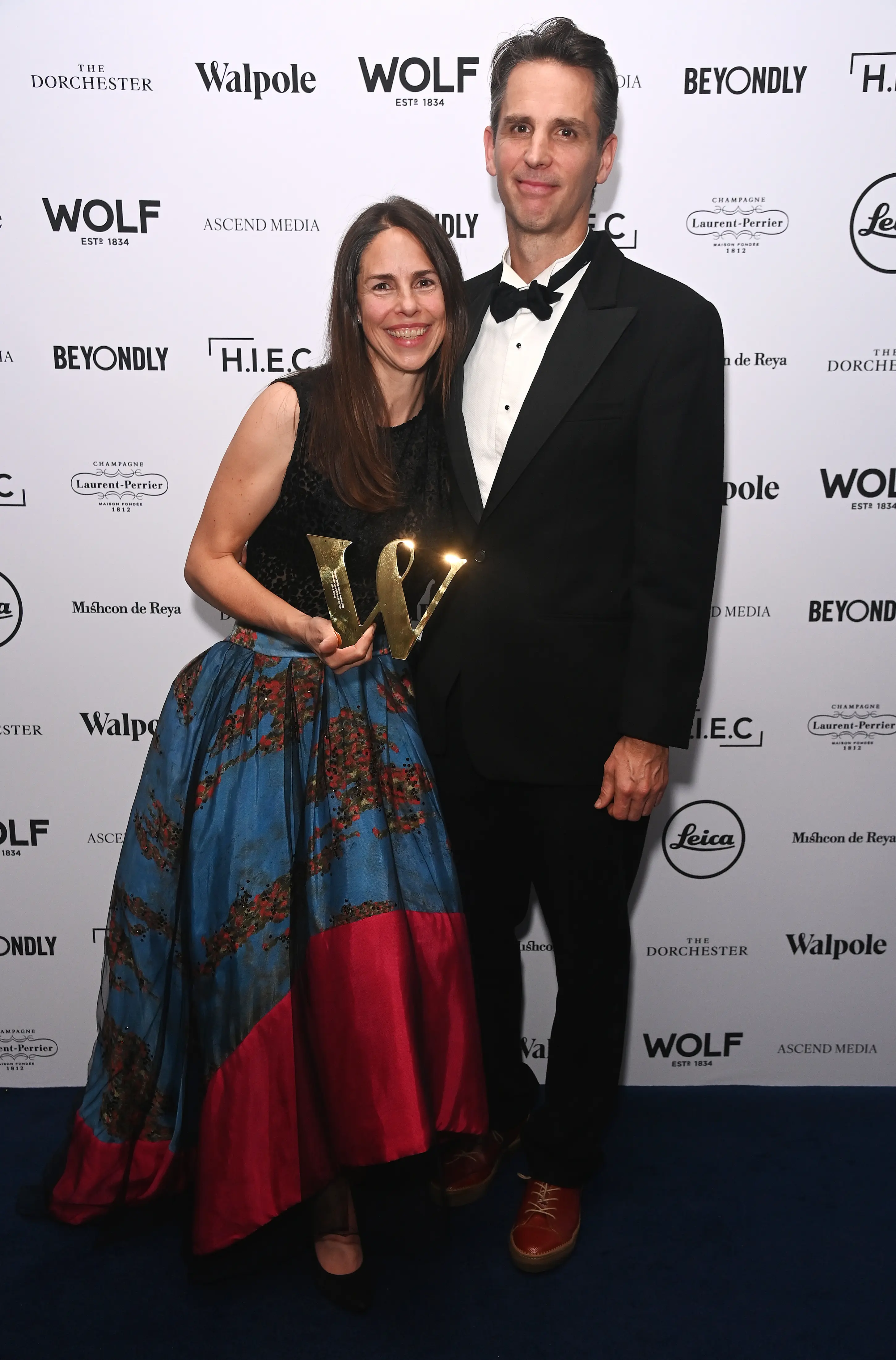 A smiling couple poses on the blue carpet at the Walpole British Luxury Awards 2025, with the woman holding a gold award trophy, standing in front of a branded step-and-repeat backdrop.