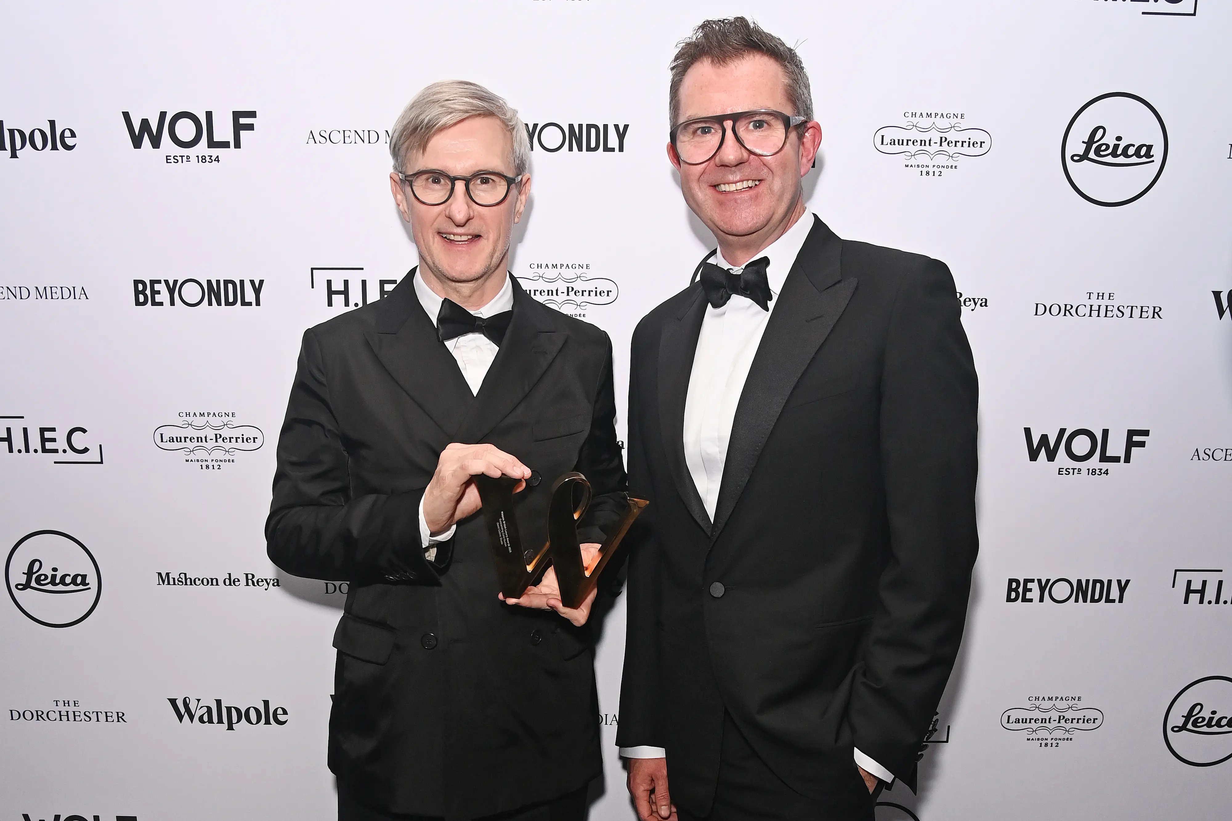 Two men in black-tie attire pose on the red carpet at the Walpole British Luxury Awards 2025, with one holding a gold award trophy in front of a branded backdrop.