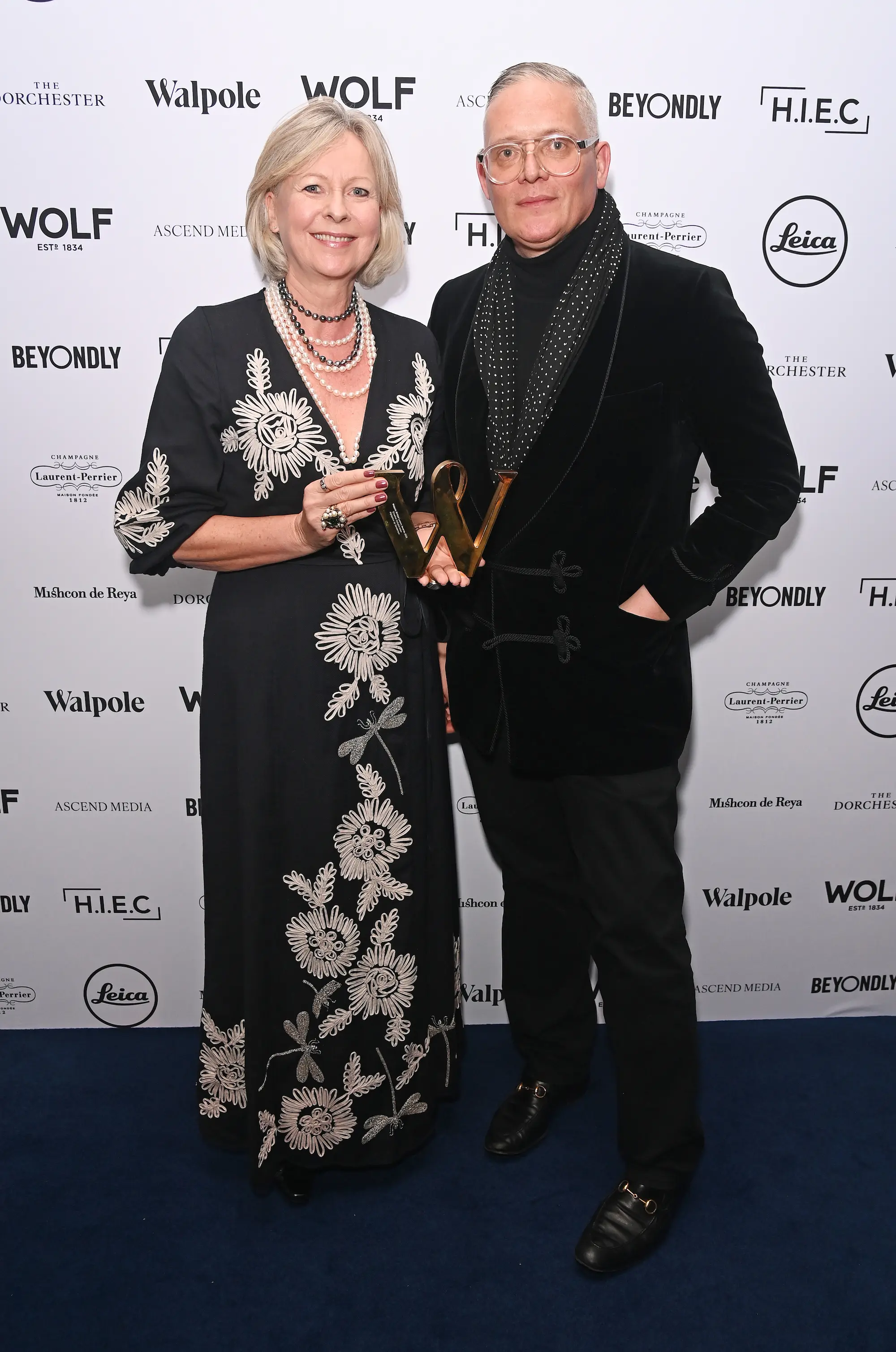 Lisa Montague and Giles Deacon pose together on the red carpet at the Walpole British Luxury Awards 2025, holding a gold award trophy in front of the event’s branded backdrop.