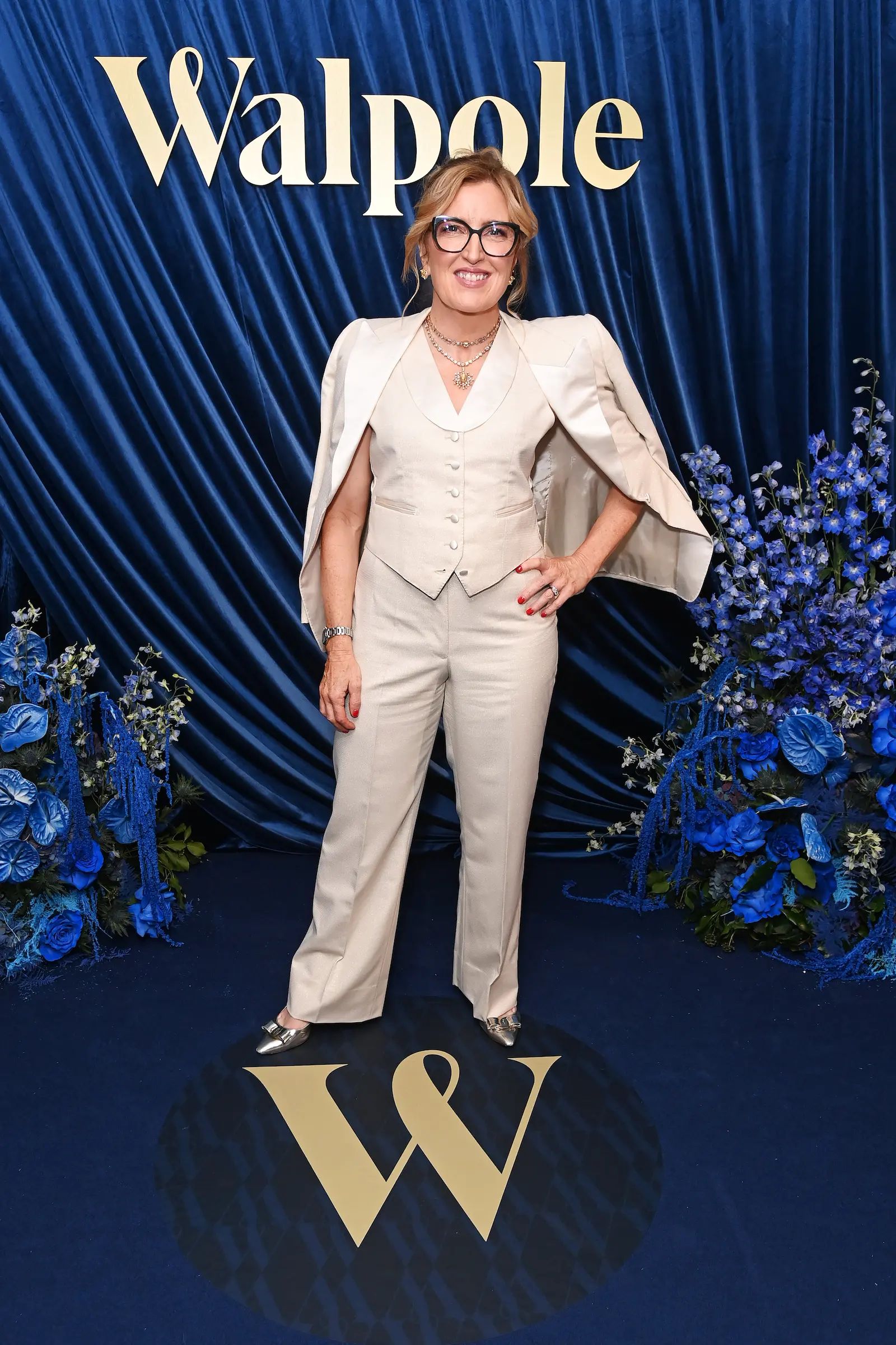Helen Brocklebank poses in a cream tailored suit against a blue velvet backdrop decorated with floral arrangements at the Walpole British Luxury Awards 2025.